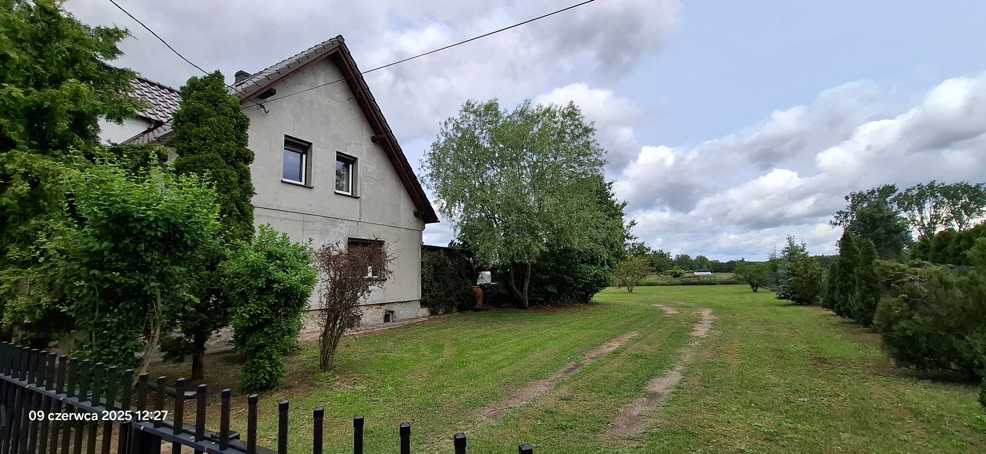 Dom piętrowy z boku, z ogrodzeniem i trawnikiem otoczony zielenią