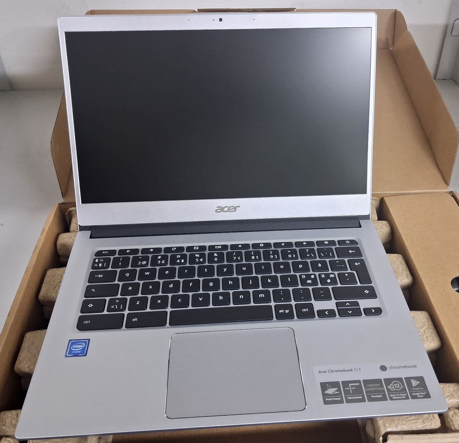 Laptop koloru srebrnego z widoczną klawiaturą oraz ekranem w opakowaniu kartonowym koloru brązowego.