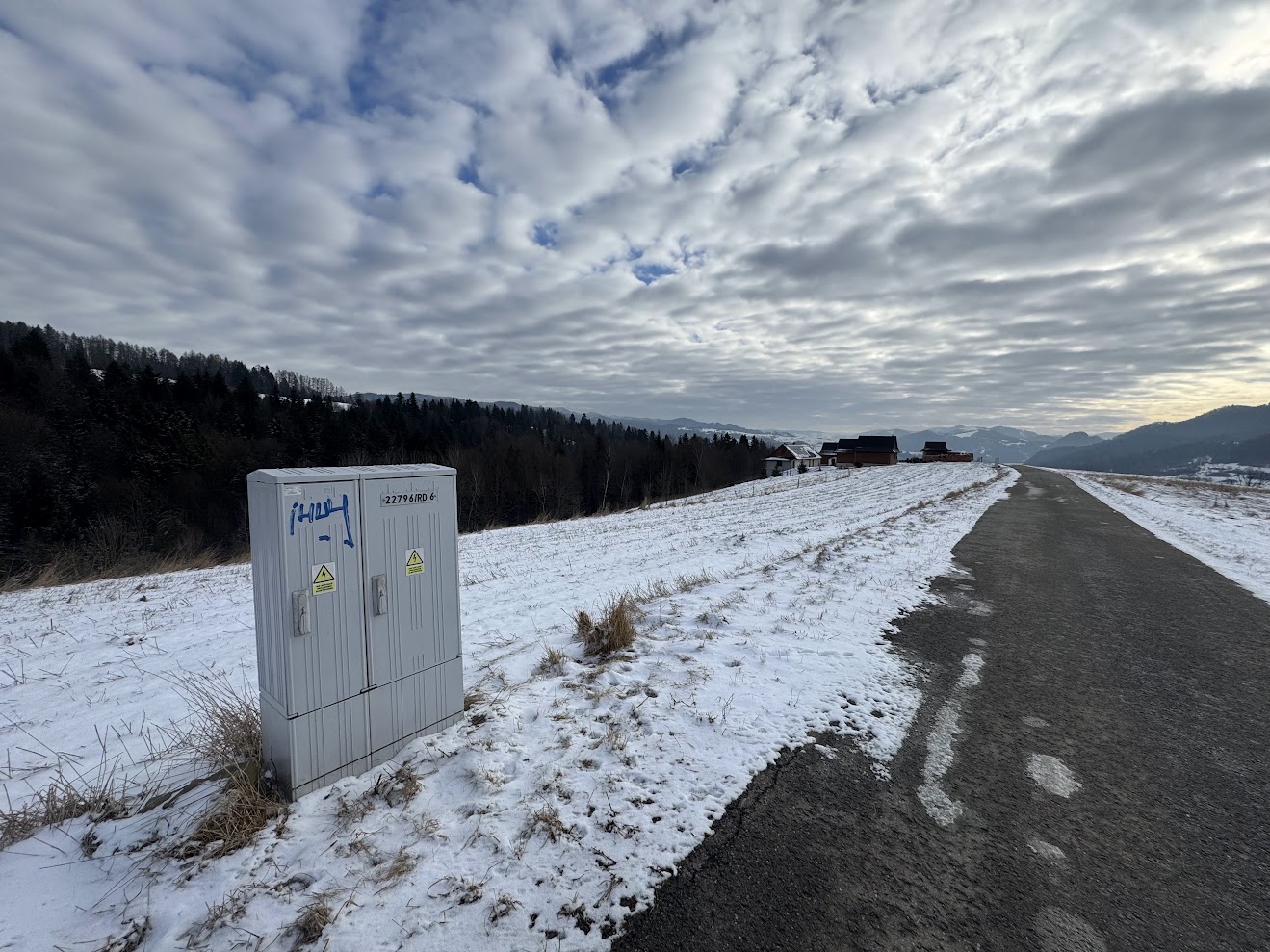 Zimowa droga biegnie przez ośnieżone wzgórza, obok skrzynki elektrycznej i zabudowań, pod rozległym, pochmurnym niebem.