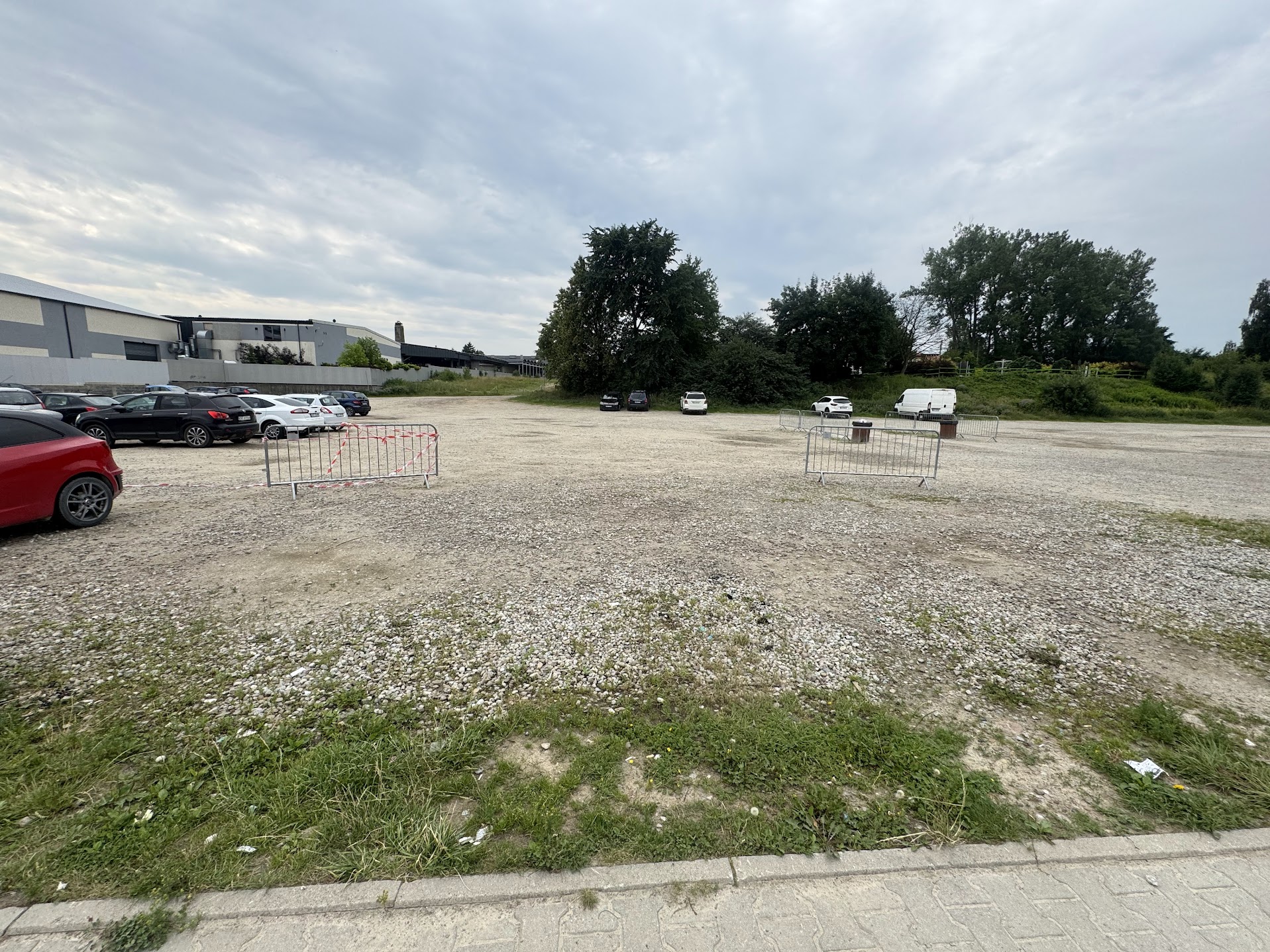 Fotografia przedstawia żwirowy plac parkingowy przy zabudowaniach przemysłowych. Na placu widoczne są samochody