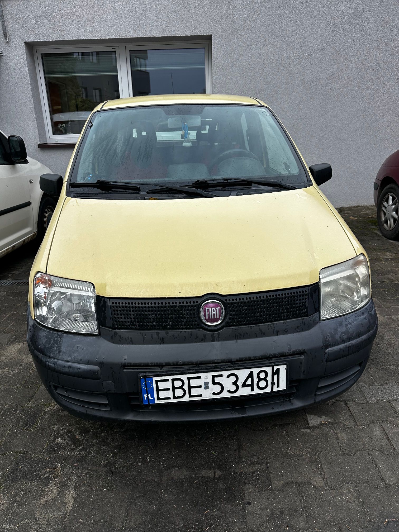 Żółty Fiat Panda II z czarnym zderzakiem i tablicą rejestracyjną EBE 53481, zaparkowany przed budynkiem.