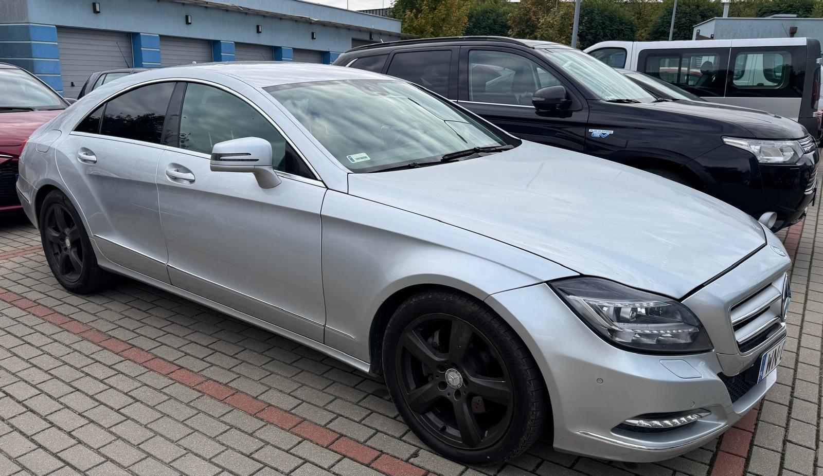 Srebrny Mercedes-Benz CLS z widocznym prawym bokiem. W tle znajdują się inne samochody. Pojazd stoi na parkingu