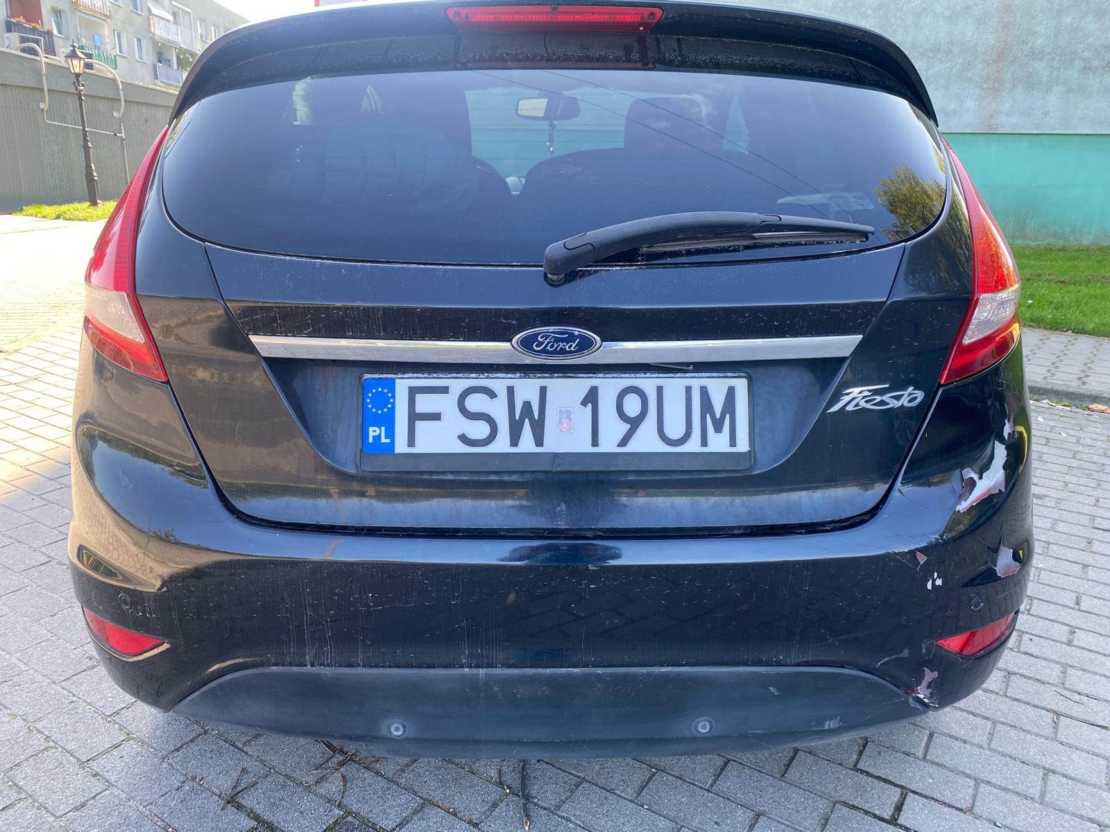 Tył Forda Fiesty koloru czarnego. Zderzak koloru czarnego uszkodzony po prawej stronie z odpryskiem lakieru.