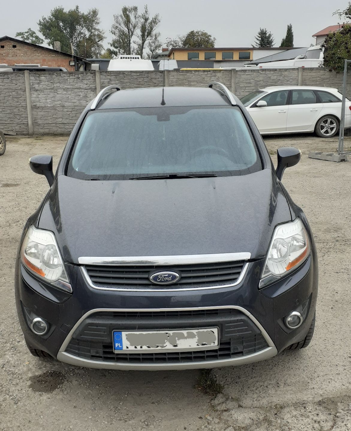 Czarny Ford Kuga pierwszej generacji, z widocznym z przodu logo marki, dużymi reflektorami i relingami dachowymi.