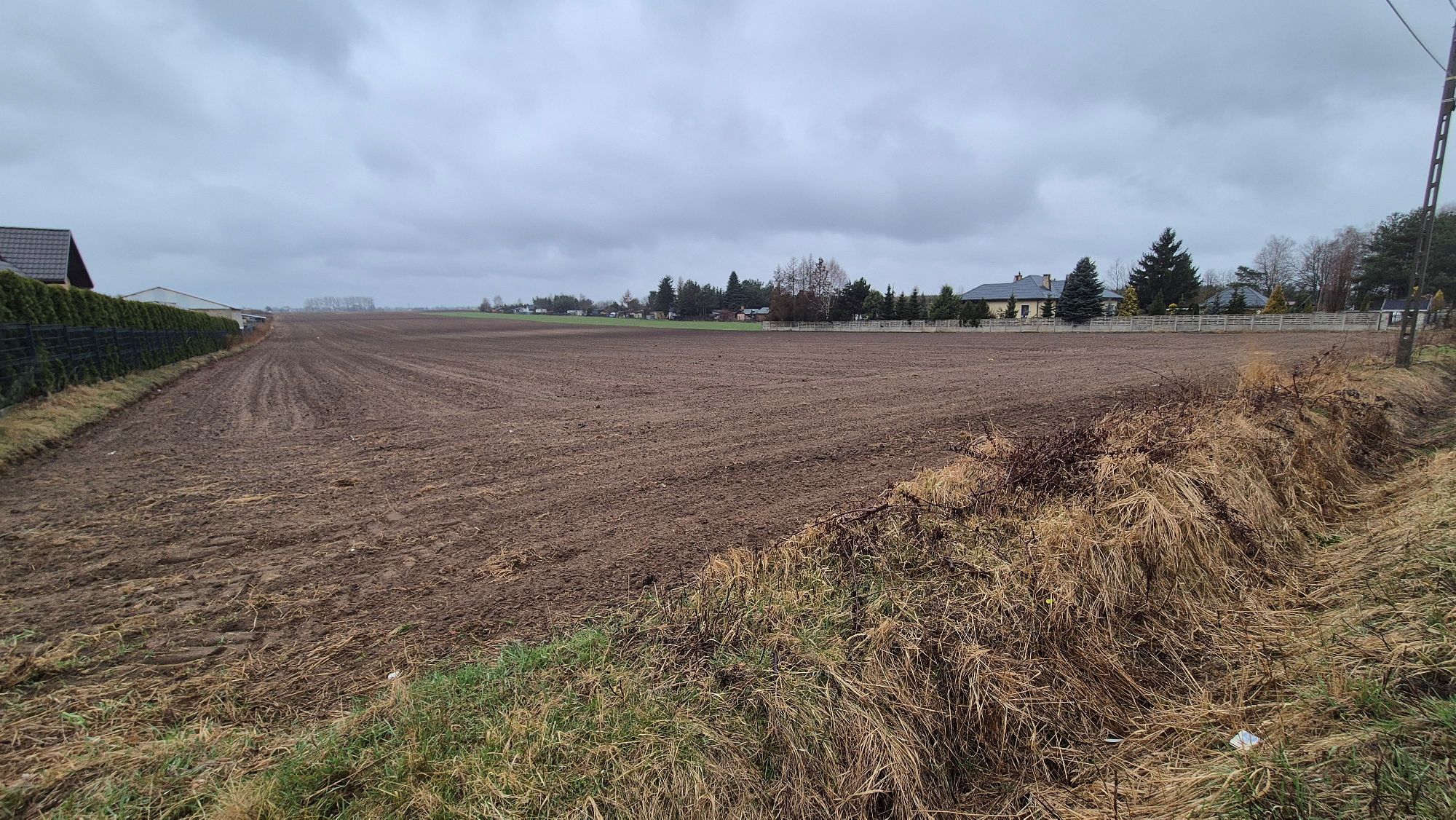 Widok na dużą, płaską działkę rolną sąsiadującą z luźną zabudową jednorodzinną i ogrodzeniami w pochmurny dzień.