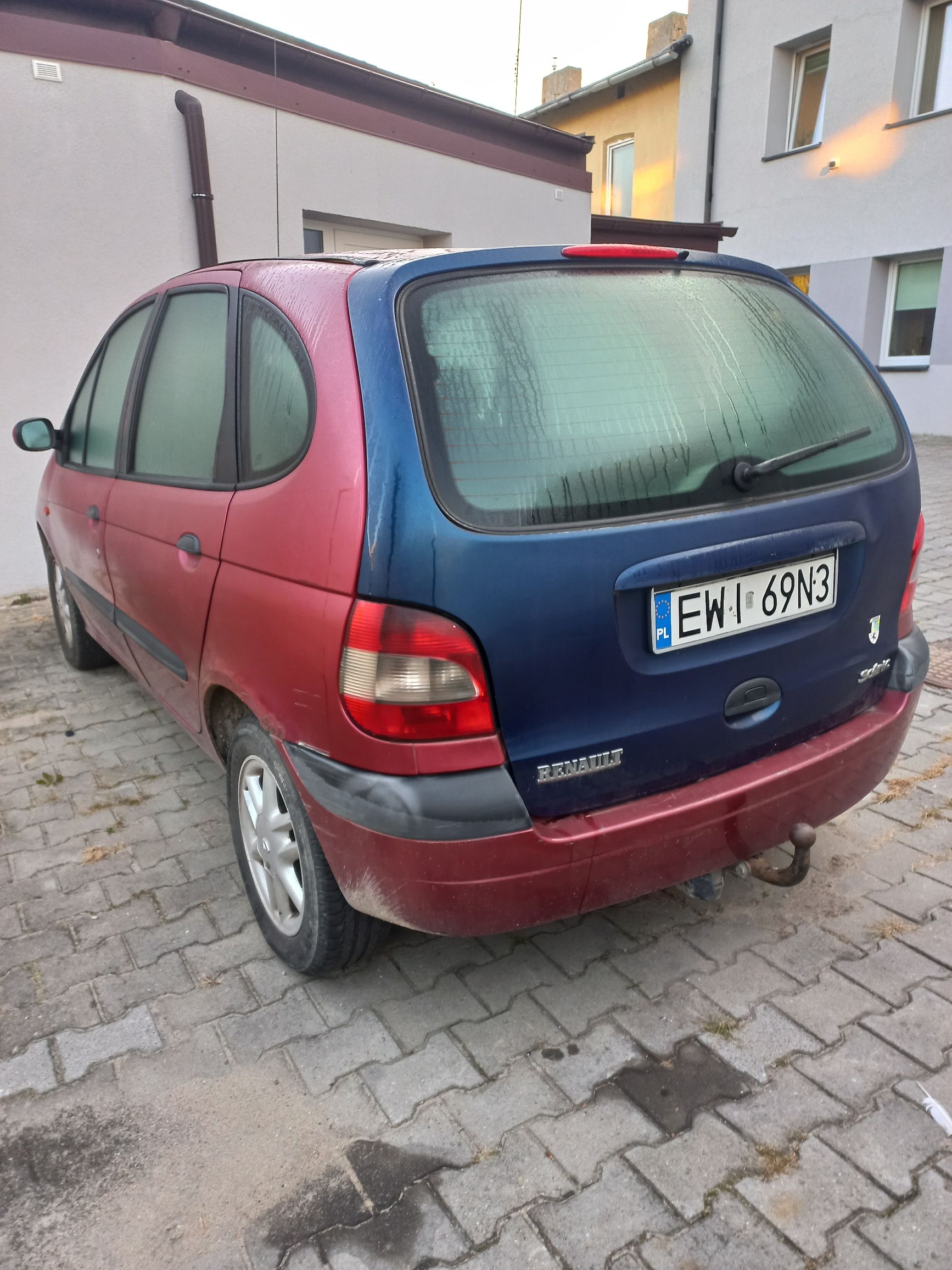 Tył czerwonego Renault Scenic z niebieską klapą bagażnika, rejestracją EWI 69N3 i zaparowanymi szybami, stojącego na kostce.