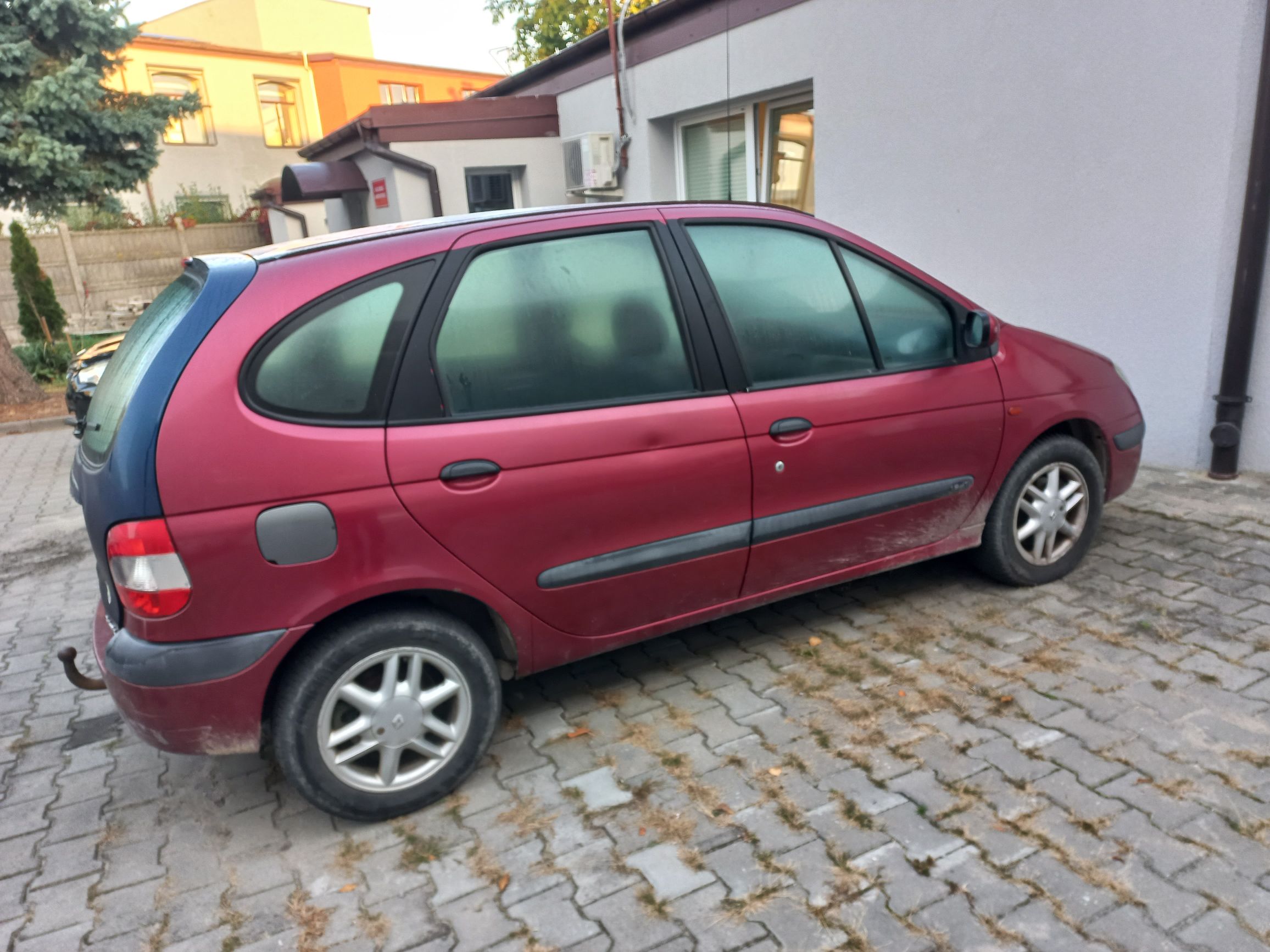 Bok czerwonego Renault Scenic z niebieskim elementem tyłu i zaparowanymi szybami, zaparkowanego na kostce brukowej.
