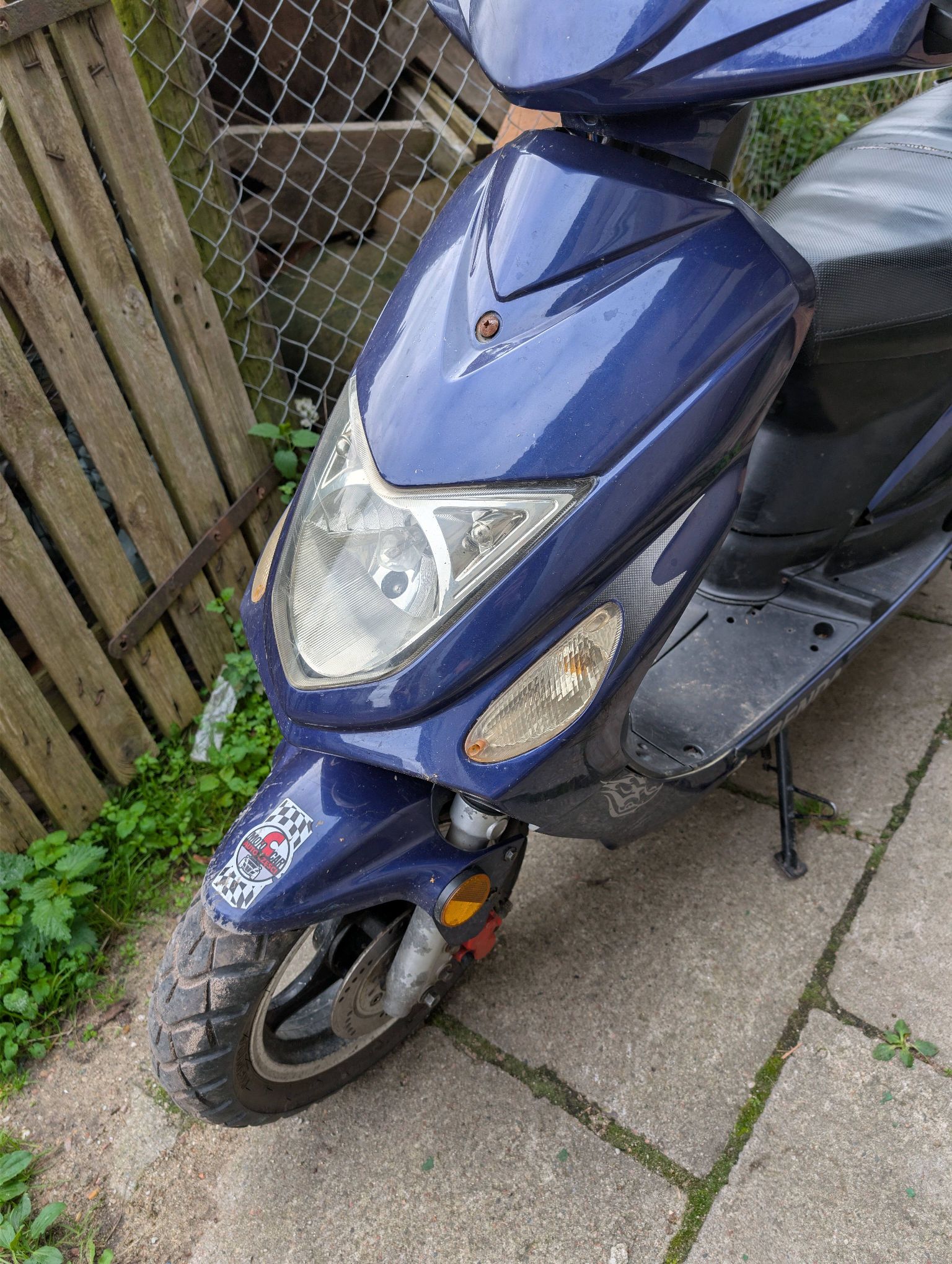 Na zdjęciu widać niebieski motorower. Na przedniej osłonie i błotniku widoczne są zabrudzenia, drobne zarysowania.