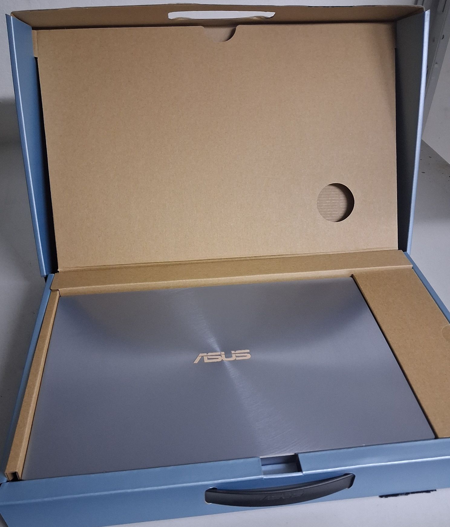 Laptop z napisem ASUS w opakowaniu kartonowym koloru niebieski-brązowego z czarnym uchwytem.
