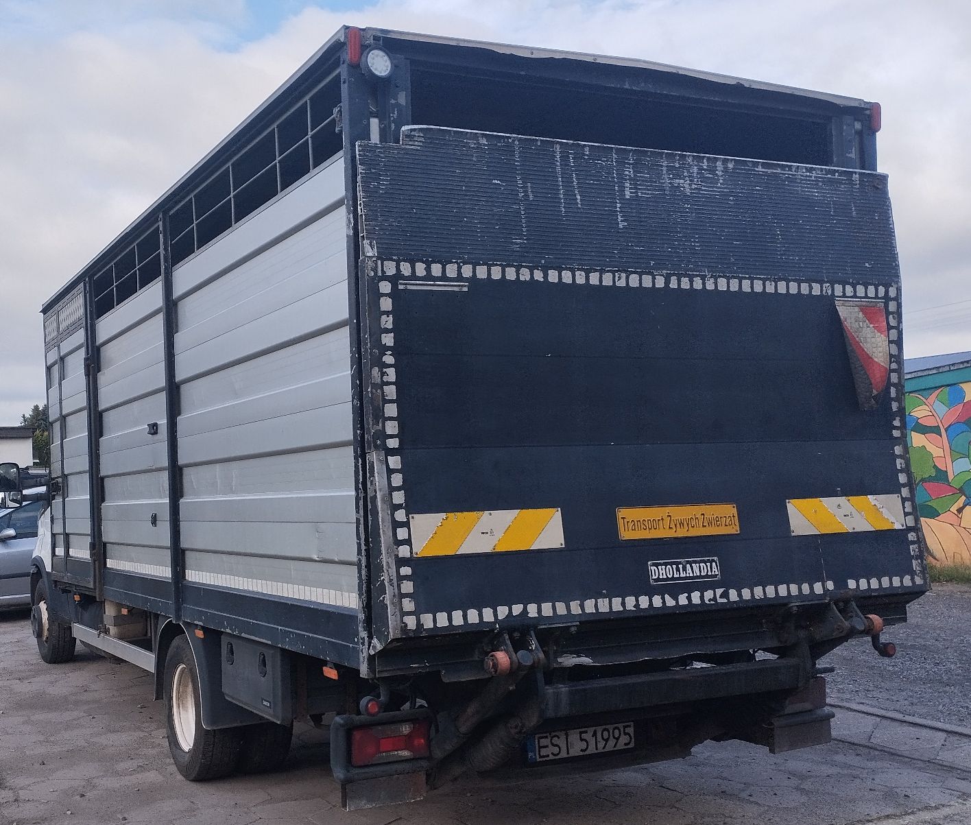 Białe Iveco Daily z lewego profilu od tyłu. Duża zabudowa do przewozu zwierząt z windą Dhollandia.