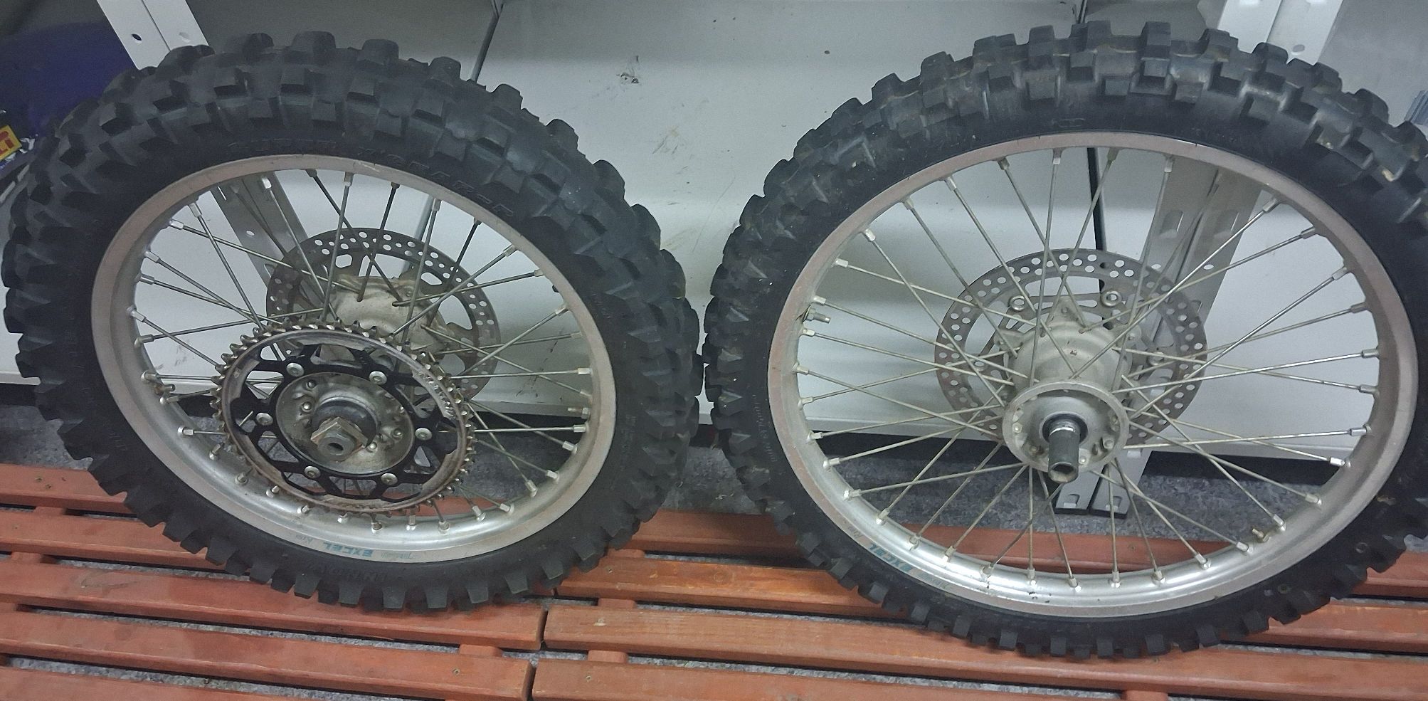 Dwa szprychowe koła motocyklowe z oponami krosowymi, tarczami hamulcowymi i zębatką napędową