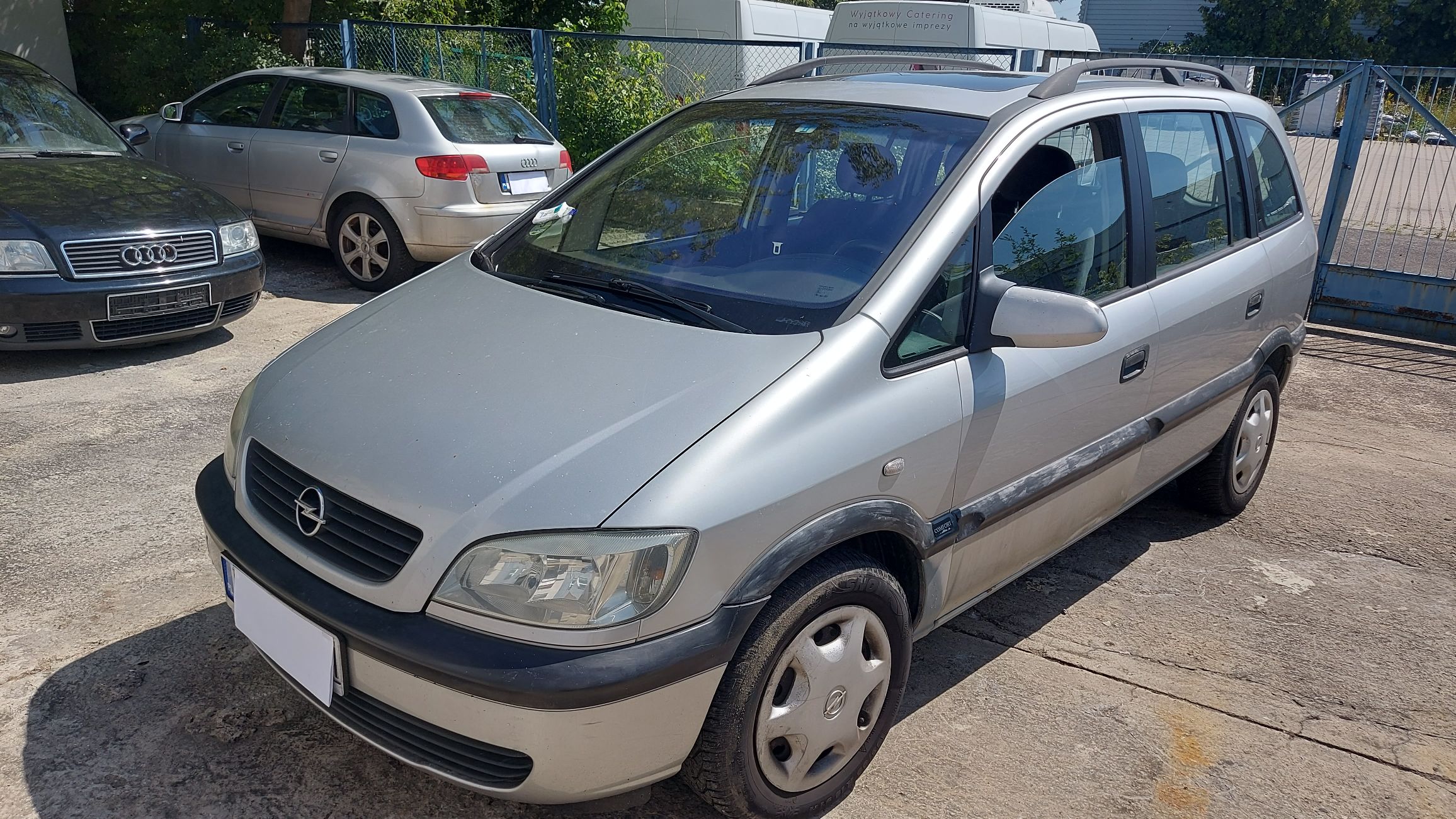 Srebrny pięciodrzwiowy Opel Zafira A zaparkowany na zewnątrz. Widok z przodu i z boku na nadwozie z relingami dachowymi.