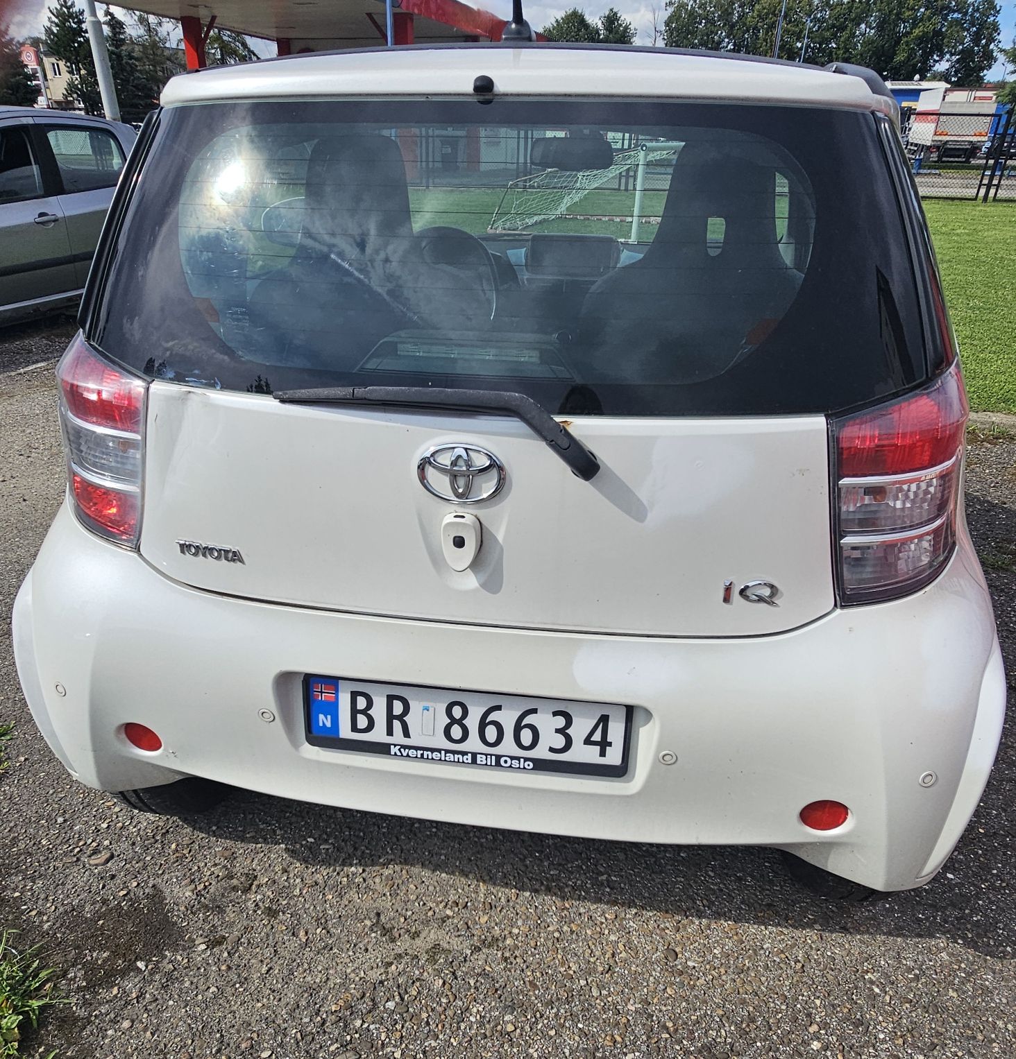 Zdjęcie przedstawia białą Toyotę iQ z norweskimi tablicami rejestracyjnymi. Pojazd ma uszkodzoną klapę bagażnika