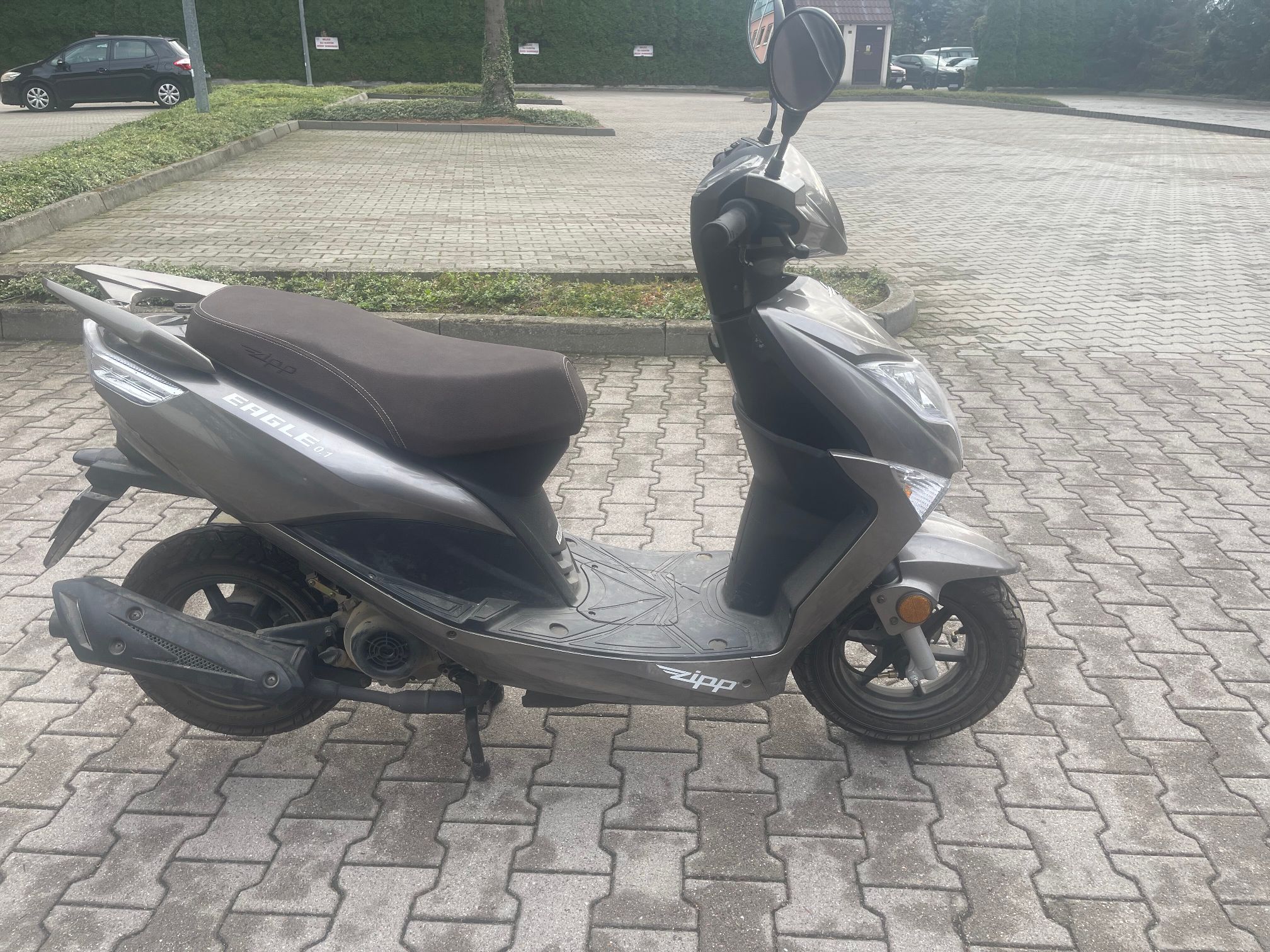 Czarny skuter. Widoczne są boczne lusterko, brązowe siedzenie, srebrne elementy nadwozia, koła, wydech oraz podpórka centralna