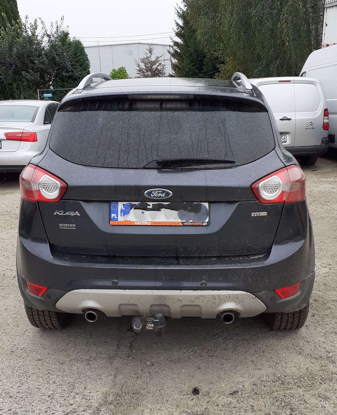 Czarny Ford Kuga widok z tyłu. Widoczna przyciemniana tylna szyba, srebrne relingi dachowe i zamontowany hak holowniczy.