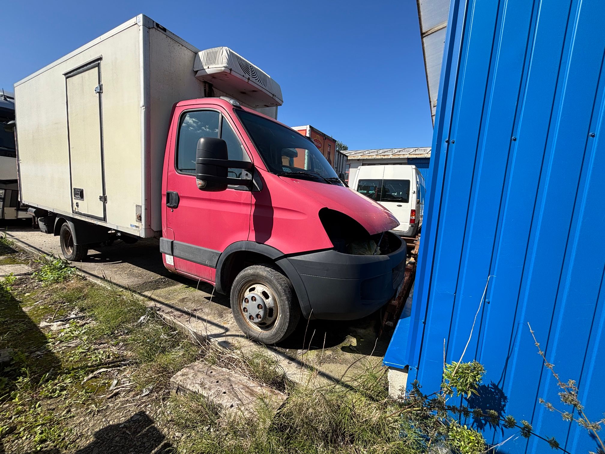 Zdjęcie przedstawia uszkodzony samochód dostawczy Iveco z białą zabudową kontenerową (chłodnią) i czerwoną kabiną kierowcy.