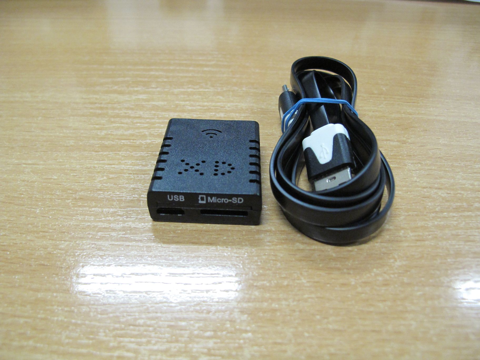 Mała, czarna mini kamera XD Micro-SD obok zwiniętego czarnego kabla USB na stole.