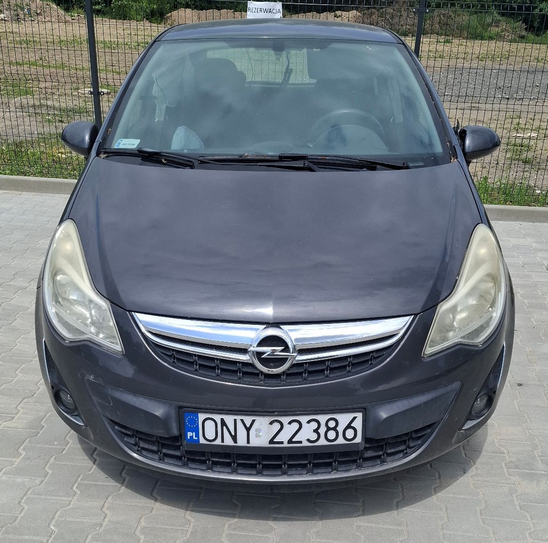 Widok z przodu grafitowego samochodu osobowego Opel Corsa. Widoczna przednia część pojazdu z reflektorami, grillem z logo Opel.