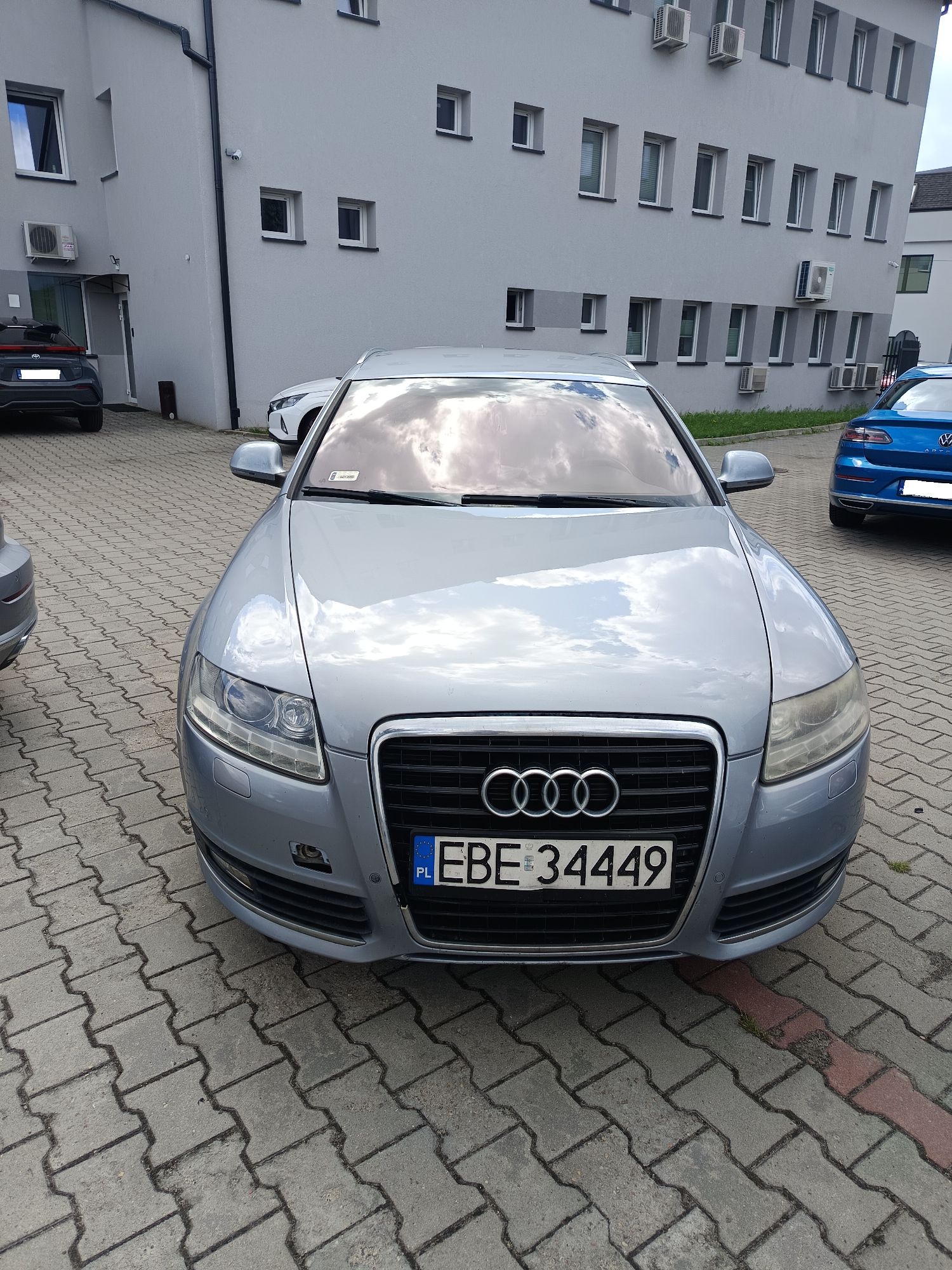 Srebrne Audi A6 zaparkowane przodem na kostce brukowej przed budynkiem.