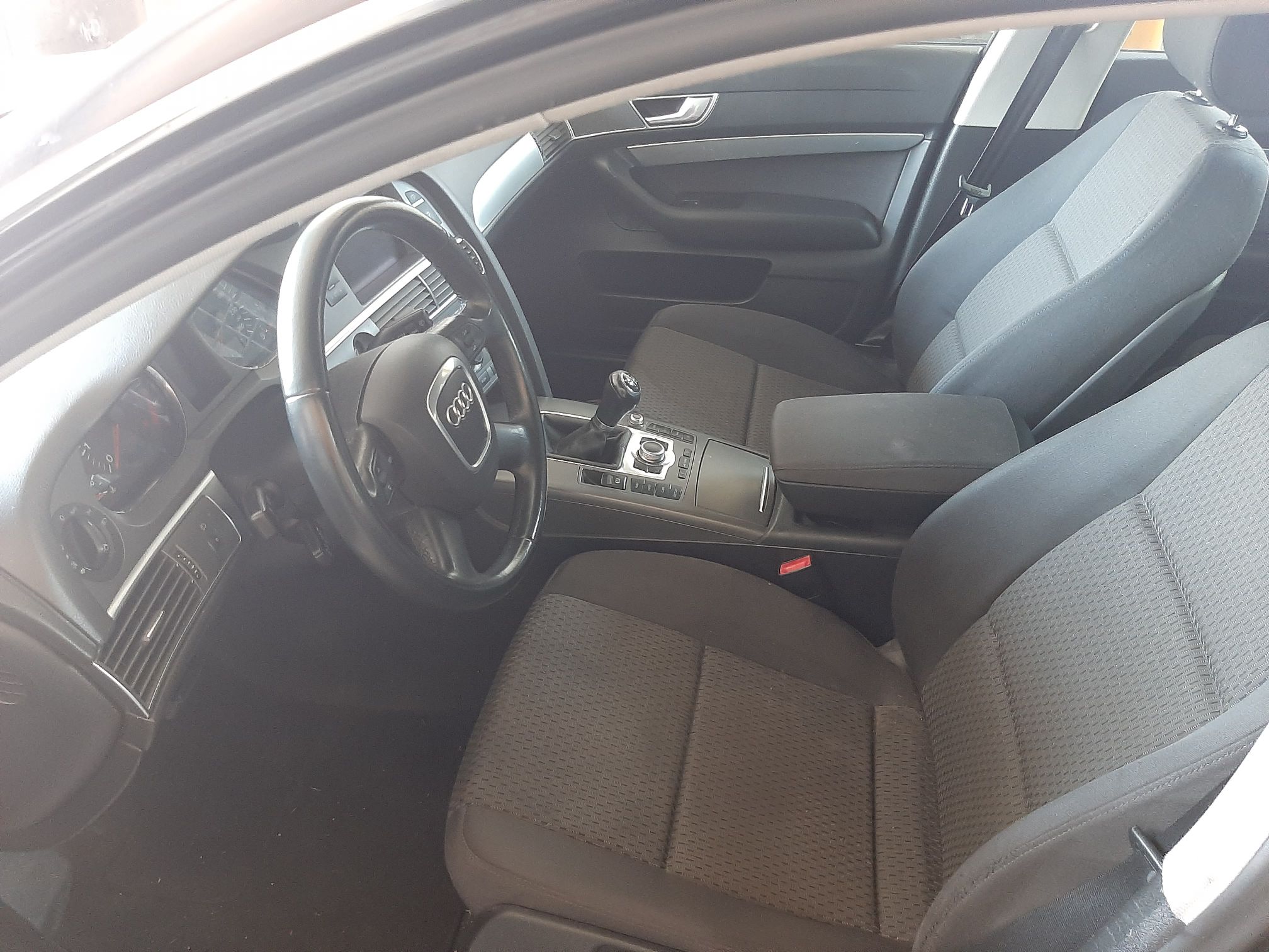 Zdjęcie przedstawia wnętrze Audi A6 z deską rozdzielczą i kierownicą. Pomiędzy fotelami jest konsola z drążkiem skrzyni biegów.
