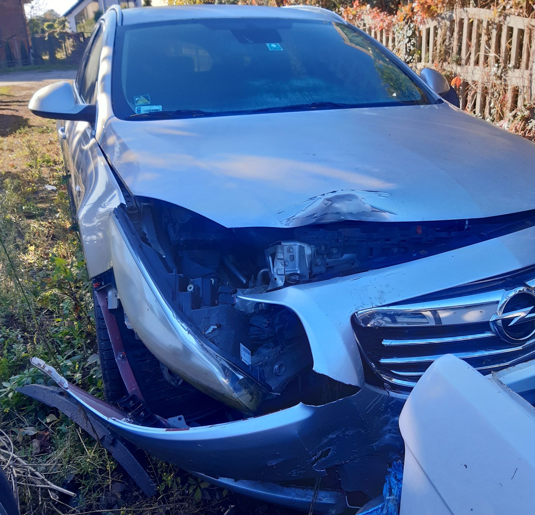 Srebrny Opel z rozbitym przodem: wgnieciona maska, oderwany zderzak i brakujący reflektor po prawej stronie.