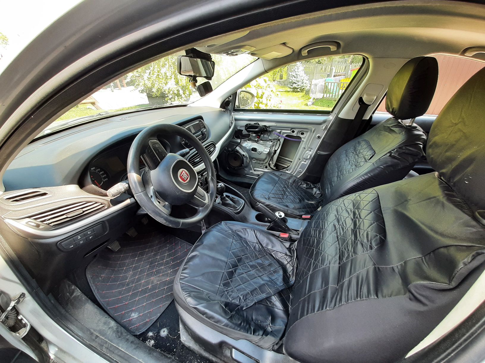 Wnętrze samochodu Fiat TIPO. Fotele są pokryte czarnymi pokrowcami. W prawych przednich drzwiach brakuje tapicerki.