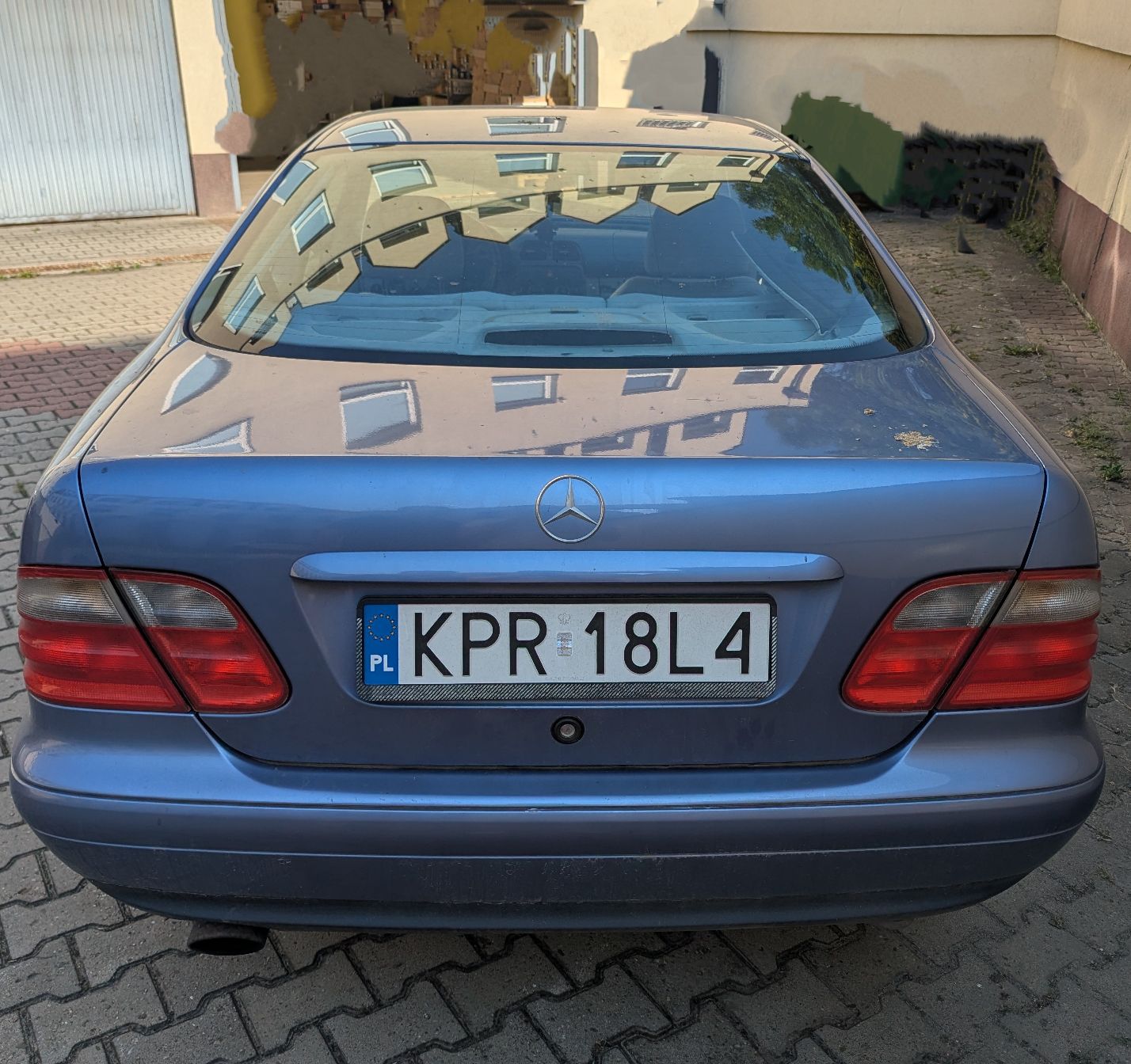 Tył samochodu Mercedes-Benz CLK 200. Szarobłękitny lakier w dobrym stanie. W centralnej części klapy bagażnika znaczek Mercedes.