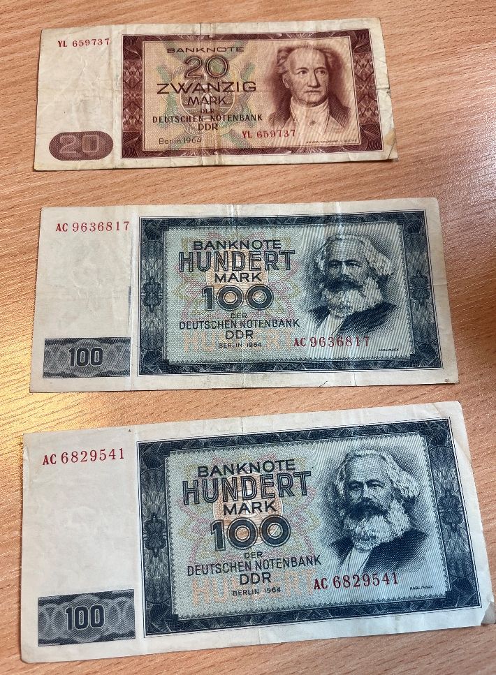 Trzy banknoty NRD z 1964 roku: jeden o nominale 20 marek i dwa o nominale 100 marek, ułożone pionowo jeden pod drugim.