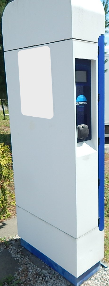 Stacja ładowania pojazdów elektrycznych marki AUTOMOC w formie wysokiego, białego słupka z gniazdem ładowania na bocznej ścianie