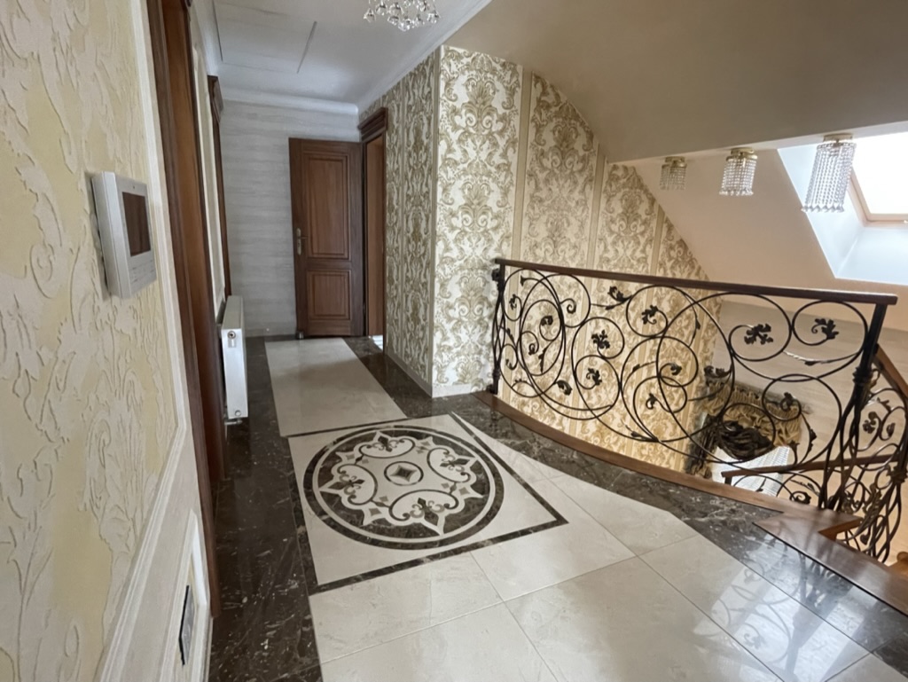 Korytarz z dekoracyjną podłogą z białych i ciemnych płytek oraz złoto-beżową tapetą, z ozdobną balustradą przy schodach.