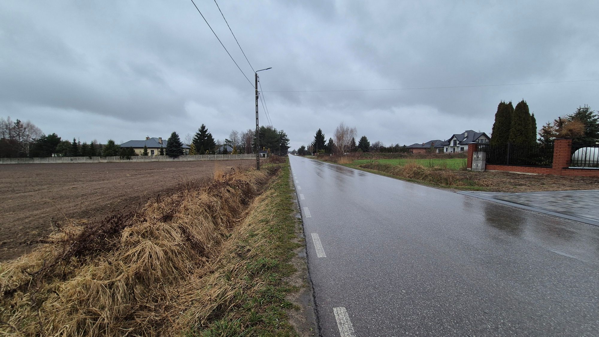 Asfaltowa droga w deszczowy dzień. Po lewej stronie widać zaorane pole, a po prawej zabudowania mieszkalne z ogrodzeniami.