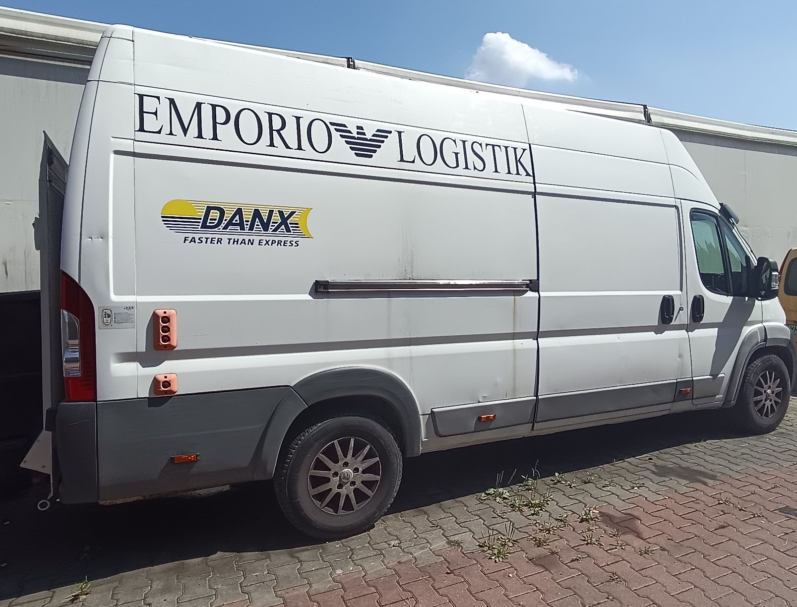 Biały samochód dostawczy widok z boku. Widoczne przesuwne drzwi boczne, na zabudowie napisy Emporio Logistic oraz logo DANX