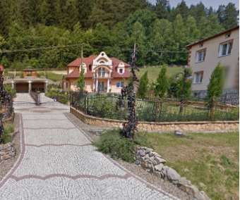 Droga wyłożona kostką prowadzi do ogrodzonego domu otoczonego zielenią i drzewami.