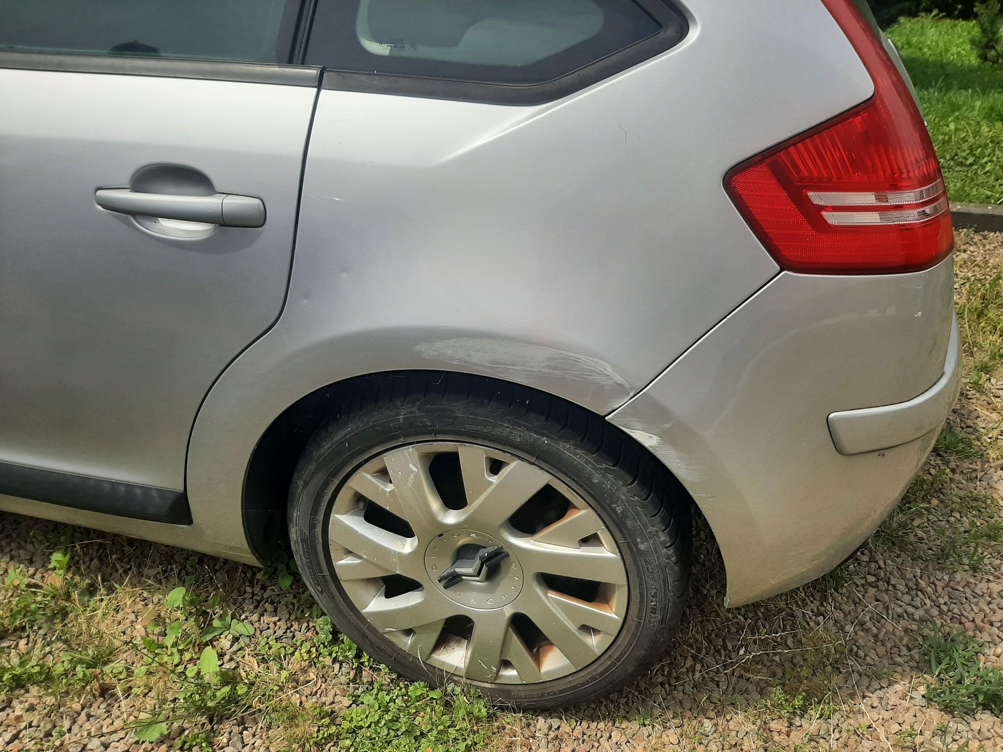 Srebrny Citroen C4. Widoczne duże, białe zarysowania i otarcia lakieru na błotniku oraz zderzaku nad tylnym, prawym kołem.