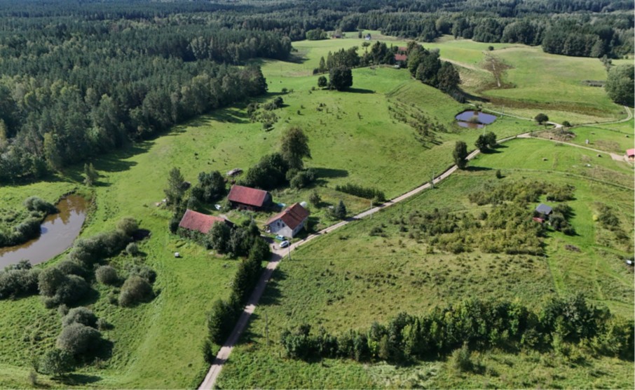 Zdjęcie przedstawia widok licytowanych działek z lotu ptaka. Znajdują się za siedliskiem, są porośnięte trawą i kilkoma drzewami