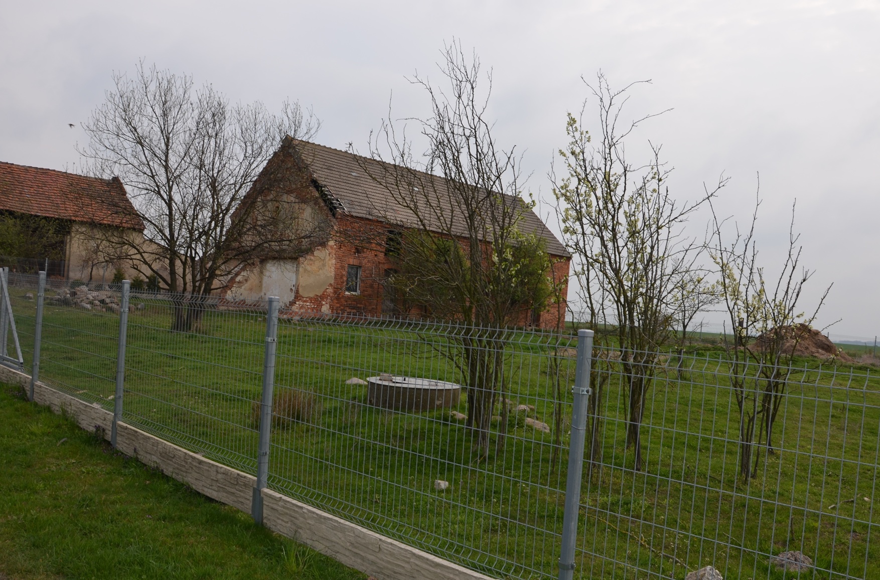 Ceglany budynek gospodarczy na ogrodzonej działce z drzewami i trawnikiem, widziany od strony drogi.