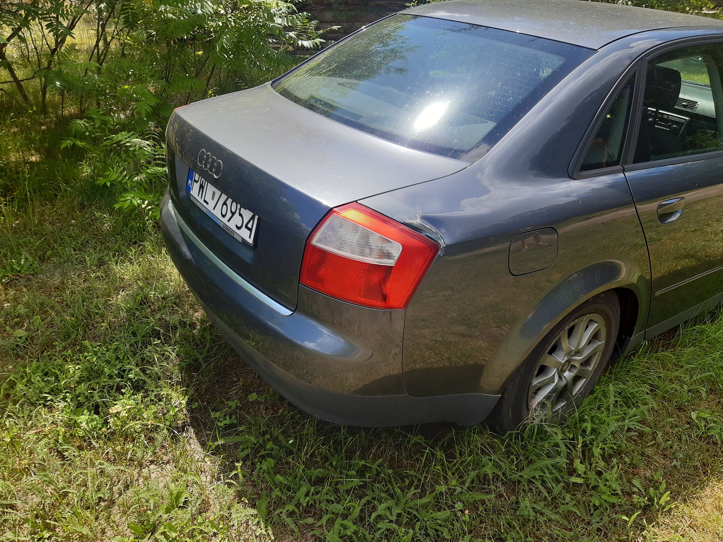 Tył szarego sedana Audi A4. Widoczne prawe tylne światło, a nad nim wgniecenie oraz zarysowanie karoserii z odpryskiem lakieru.