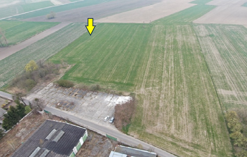 Fotografia przedstawia pola uprawne, drogę, zabudowania gospodarcze i zadrzewienia. Żółta strzałka wskazuje fragment pola.