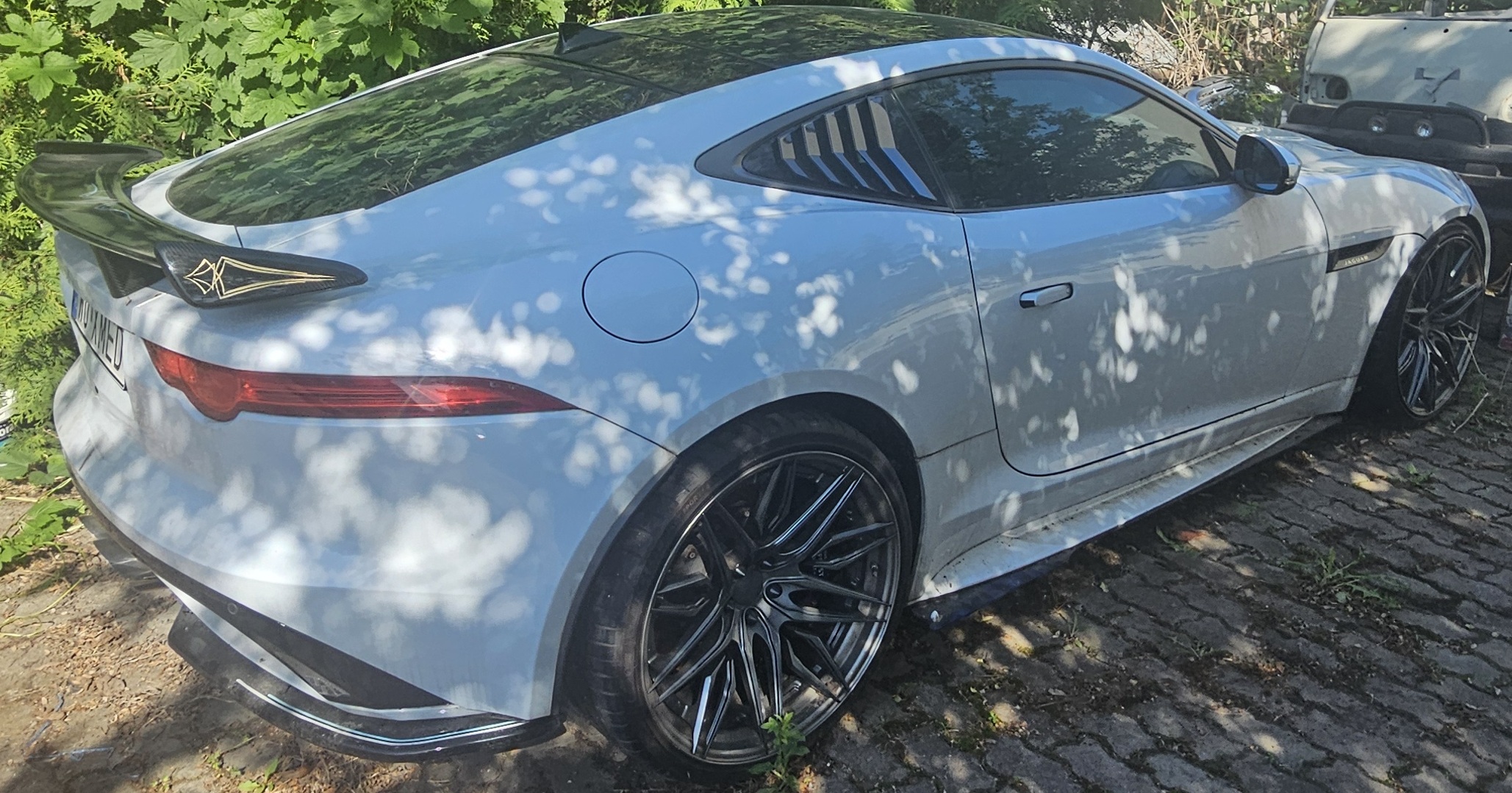 Zdjęcie przedstawia prawy tylny bok białego auta. Na pierwszym planie widoczna jest duża aluminiowa felga.