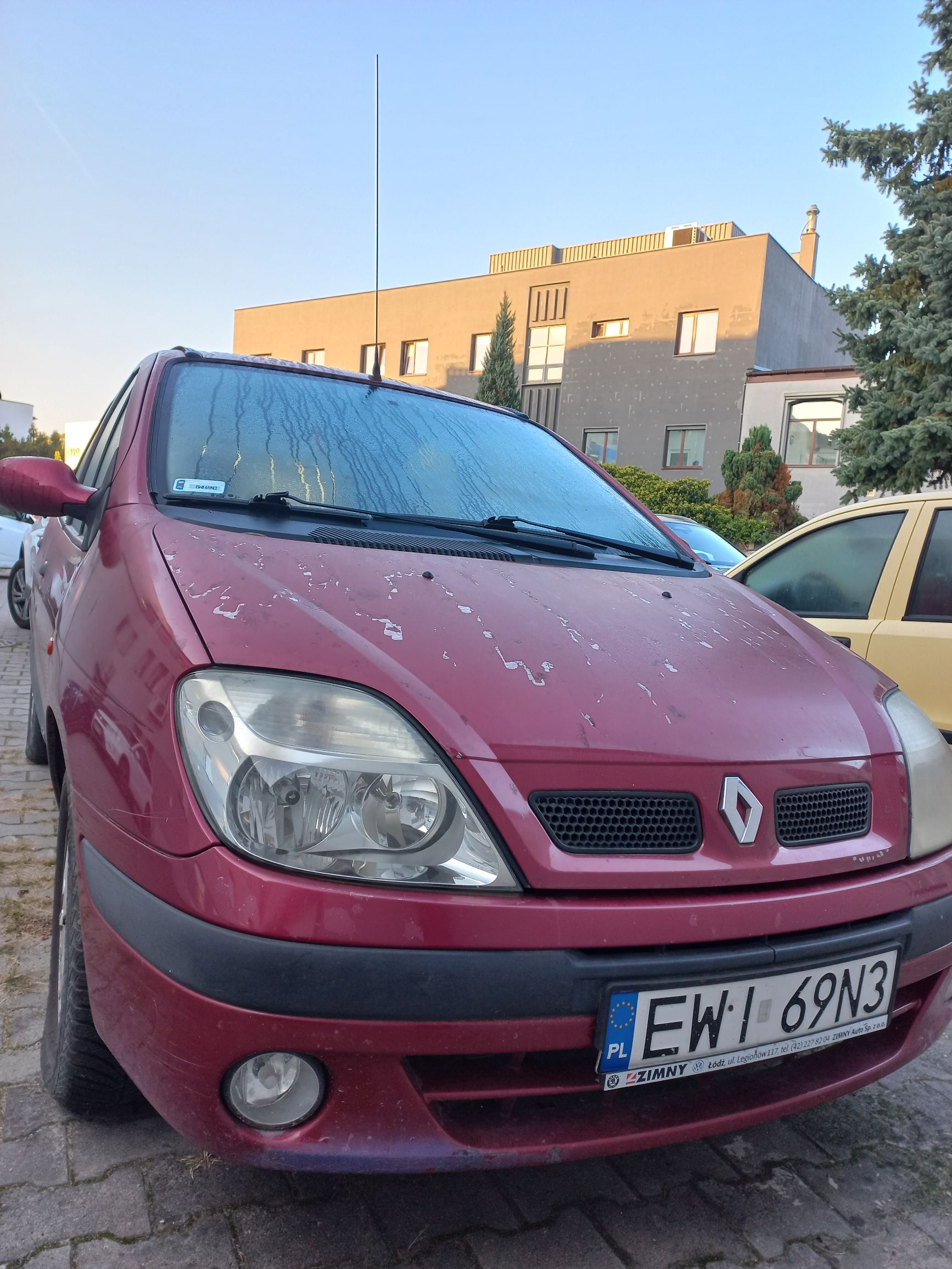 Przód czerwonego samochodu marki Renault zaparkowanego na brukowanym parkingu przed nowoczesnym budynkiem.