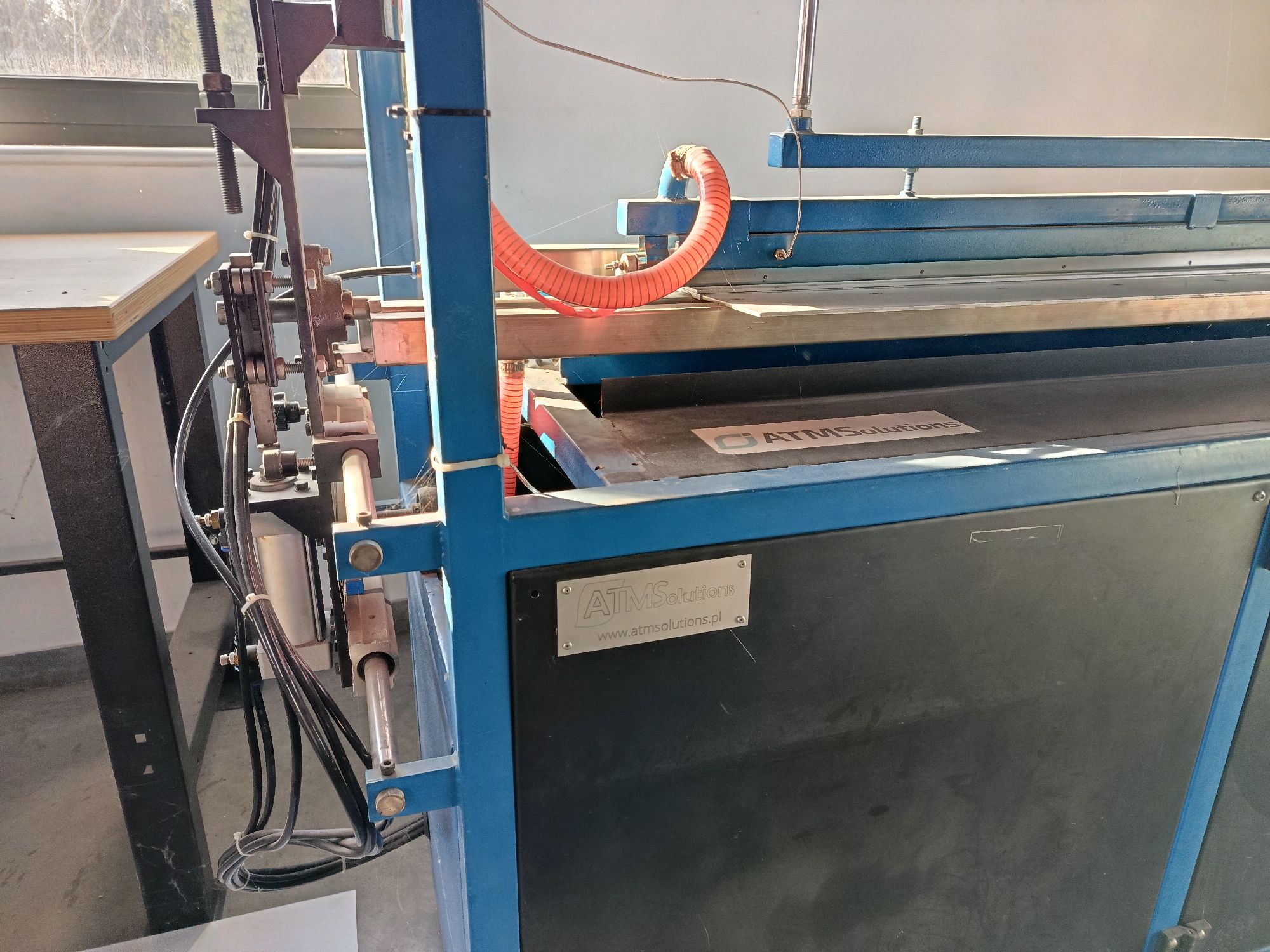 Duża, przemysłowa maszyna produkcyjna (automatyczna giętarka) o niebieskiej metalowej konstrukcji szkieletowej.