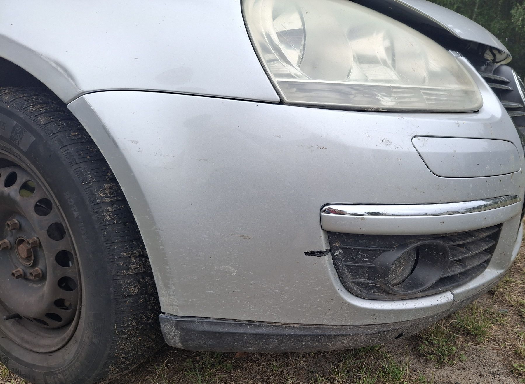 Zbliżenie na uszkodzony zderzak srebrnego Volkswagena, widoczne pęknięcie obok kratki halogenu i zmatowiona lampa.