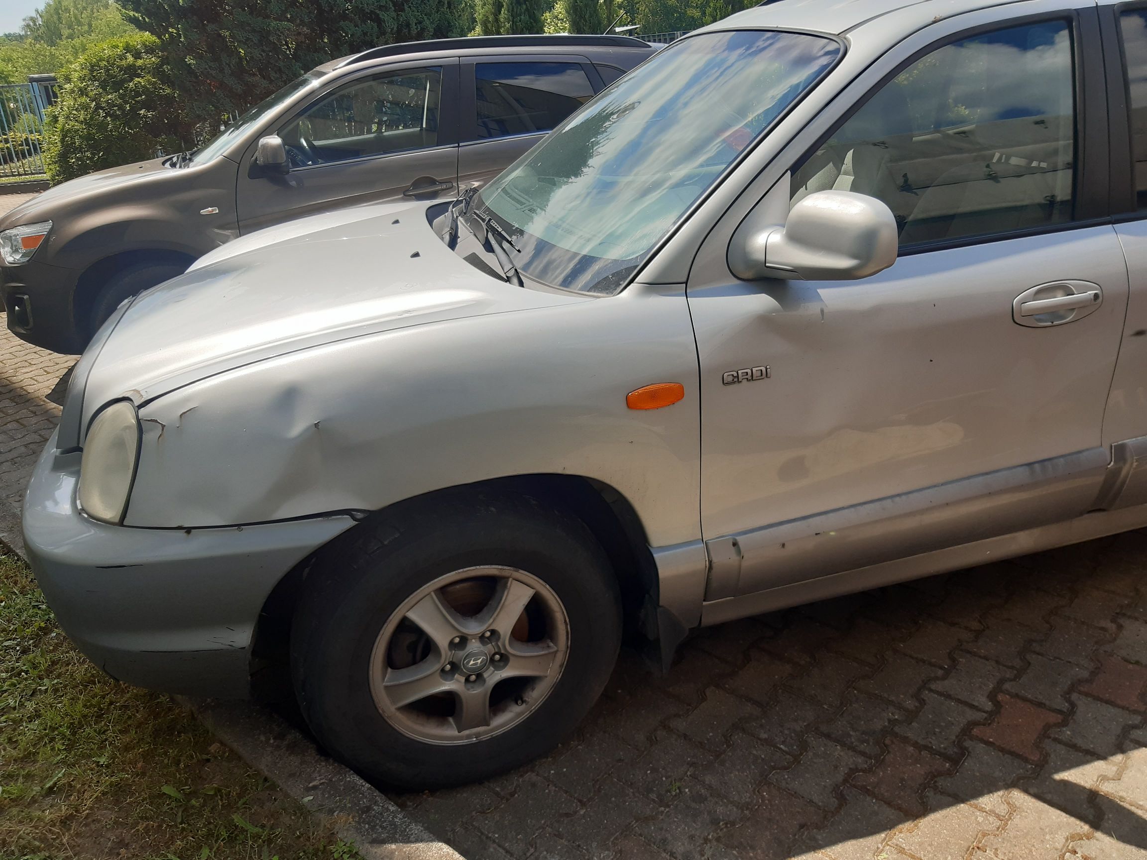 Srebrny samochód osobowy marki Hyundai model Santa Fe pierwszej generacji. Pojazd nosi wyraźne ślady uszkodzeń z przodu.
