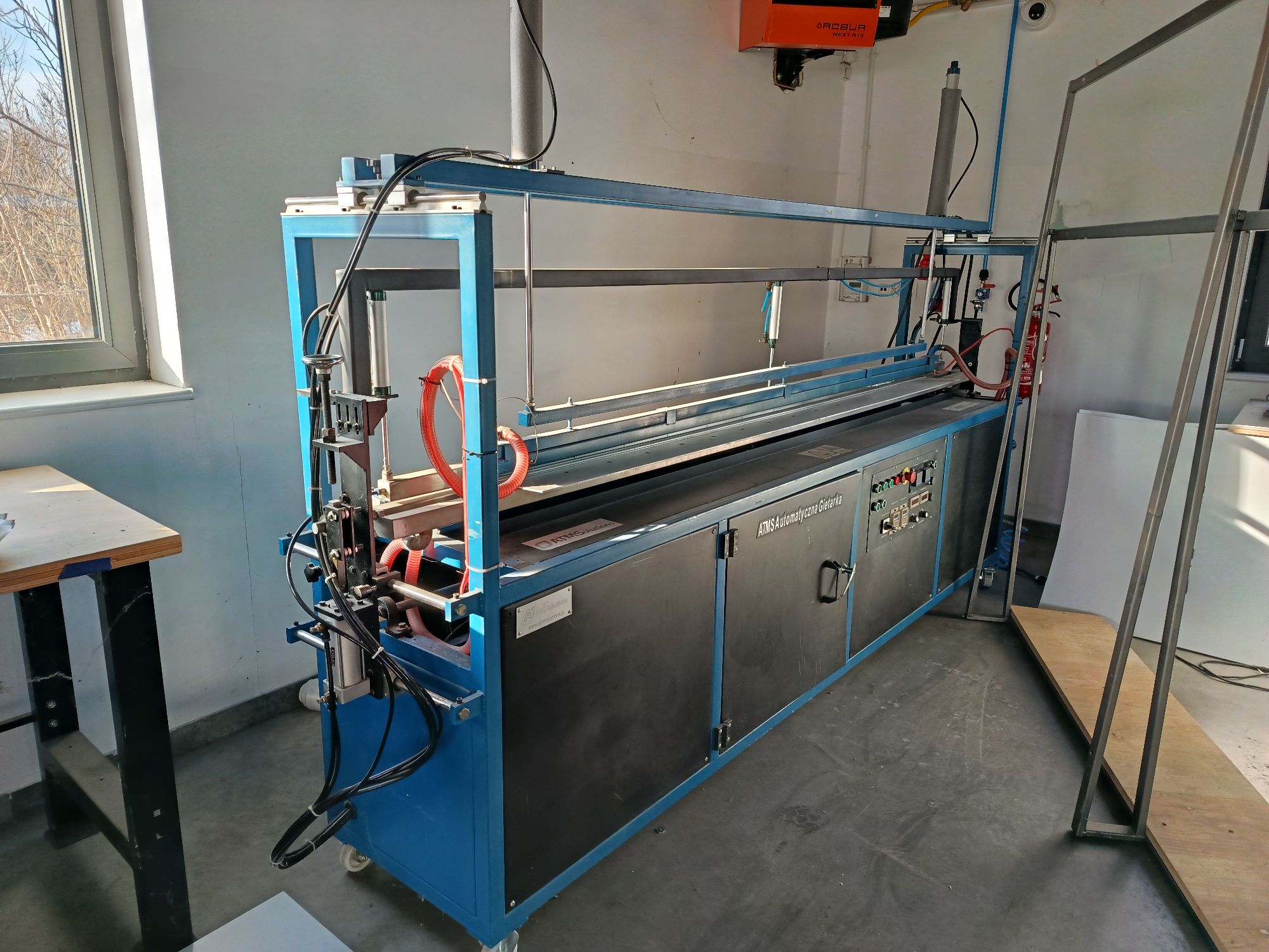 Duża, przemysłowa maszyna produkcyjna (automatyczna giętarka) o niebieskiej metalowej konstrukcji szkieletowej.