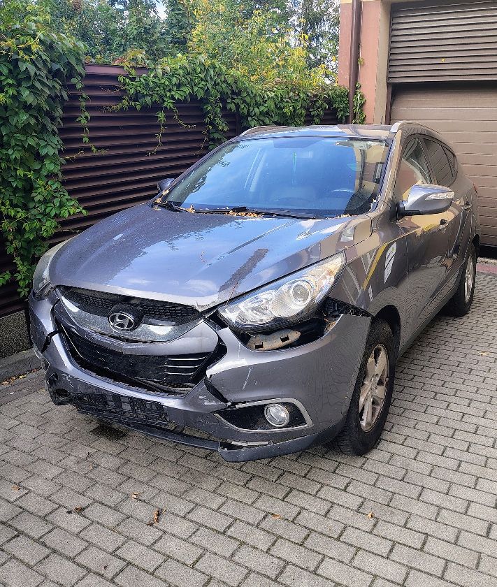 Pięciodrzwiowy szary samochód osobowy typu SUV. Uszkodzony przedni zderzak. Część maski zgięta i porysowana.