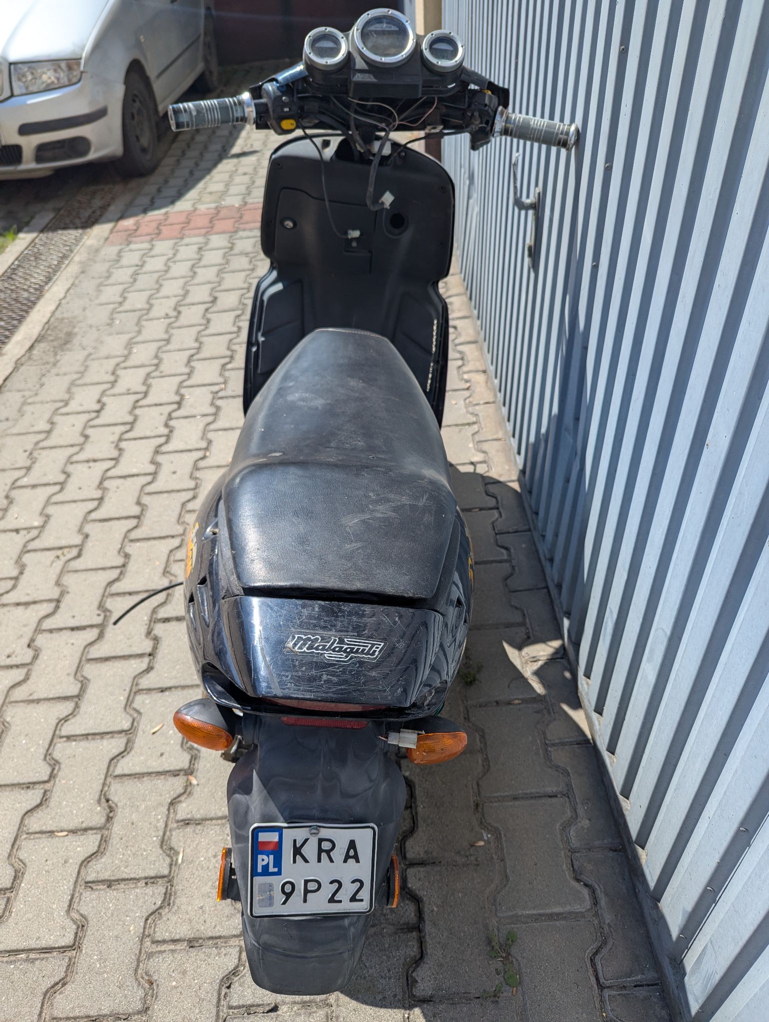 Tył czarnego motoroweru Malaguti. Kokpit kierownicy odkręcony, odsłonięte przewody elektryczne, kanapa w dobrym stanie.