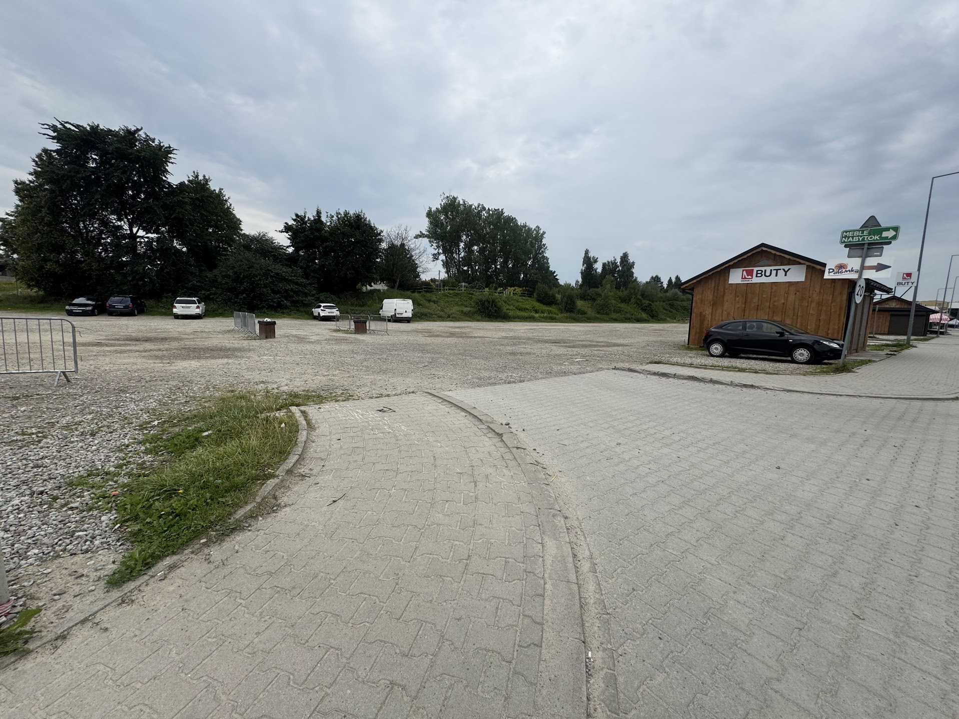 Fotografia przedstawia dojazd do niewielkiego parkingu przy budynku usługowym, z samochodami, żwirową nawierzchnią