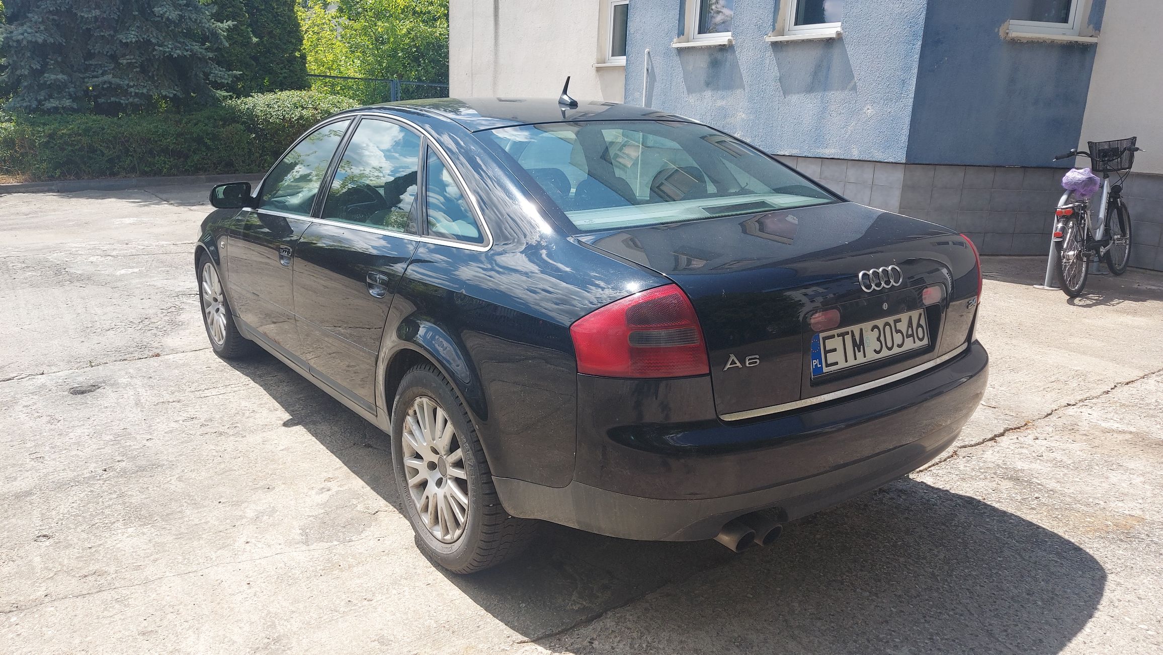 Widoczny tył i lewy bok czarnego Audi A6 C5, zaparkowanego na betonowym podjeździe obok budynku.