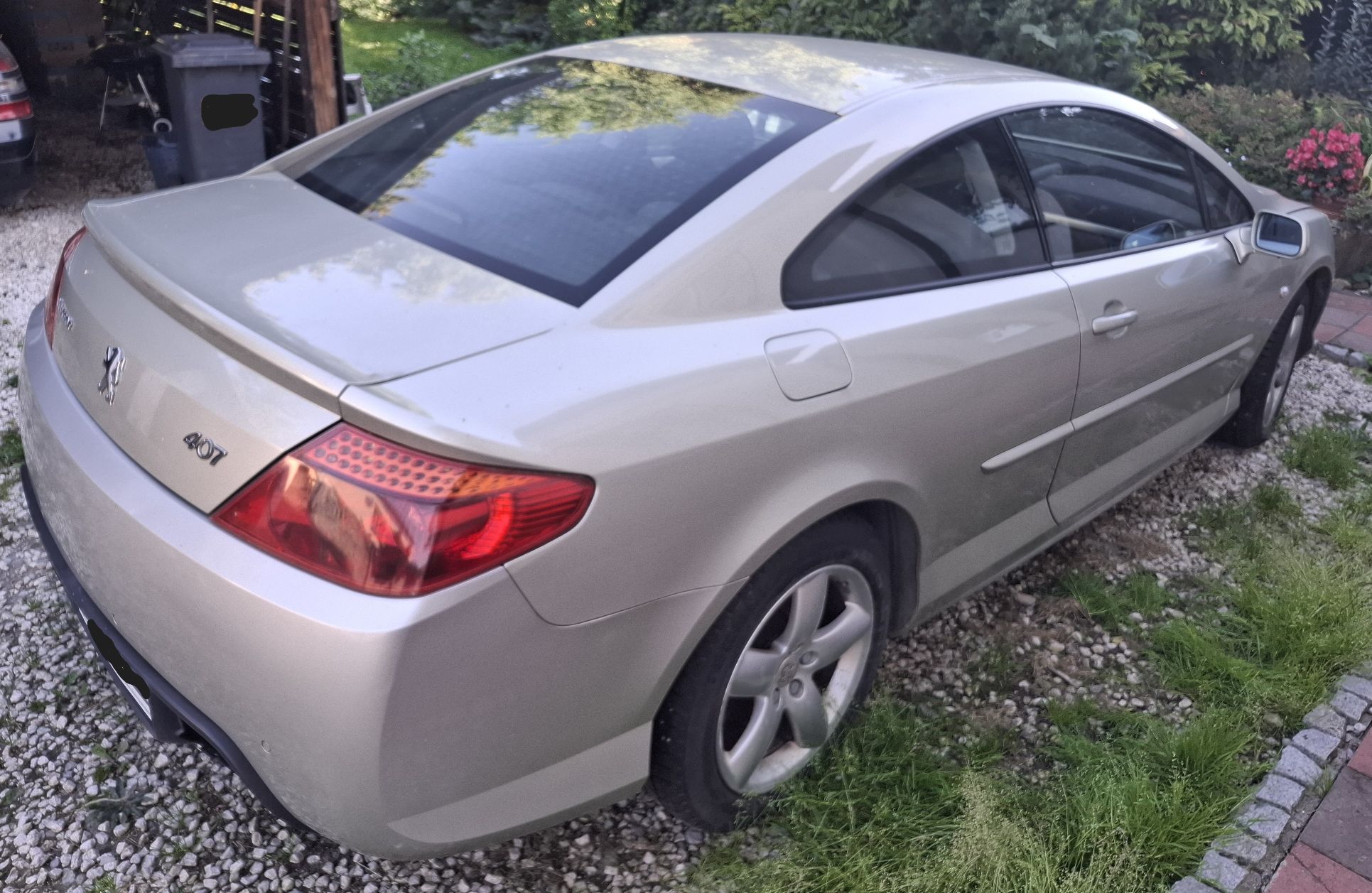 Na zdjęciu widczony jest Peugeot 407 Coupé w beżowo-popielatym kolorze nadwozia. Widok pojazdu od tyłu, z prawego boku.