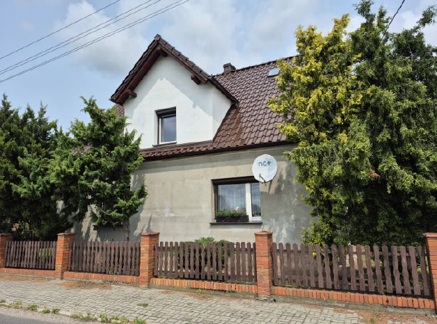 Dom ze spadzistym brązowym dachem, otoczony drewnianym płotem i zielenią